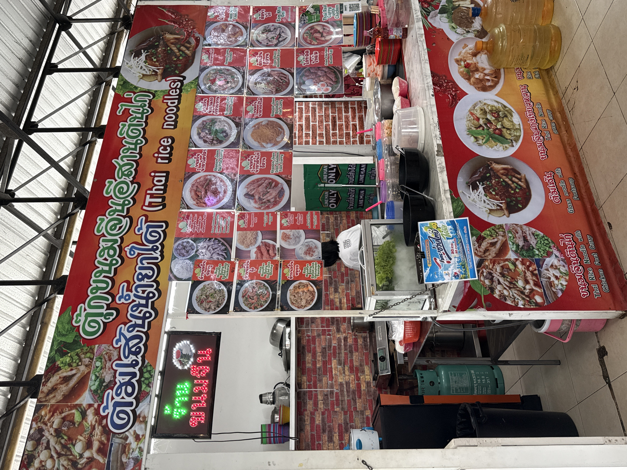 The image shows an outdoor food stall in Baan Bo Phut, Thailand. The stall features a red banner with Thai text and "Thai rice noodles" in English, displaying images of various noodle dishes. A woman with dark hair, wearing a white t-shirt, is visible behind the counter, facing away from the camera. The stall is stocked with ingredients and cooking equipment, including pots, bowls, and bottles of liquid. In the foreground, several large water jugs are placed on the counter. The environment appears to be a bustling market or street food area, with a metal roof overhead.