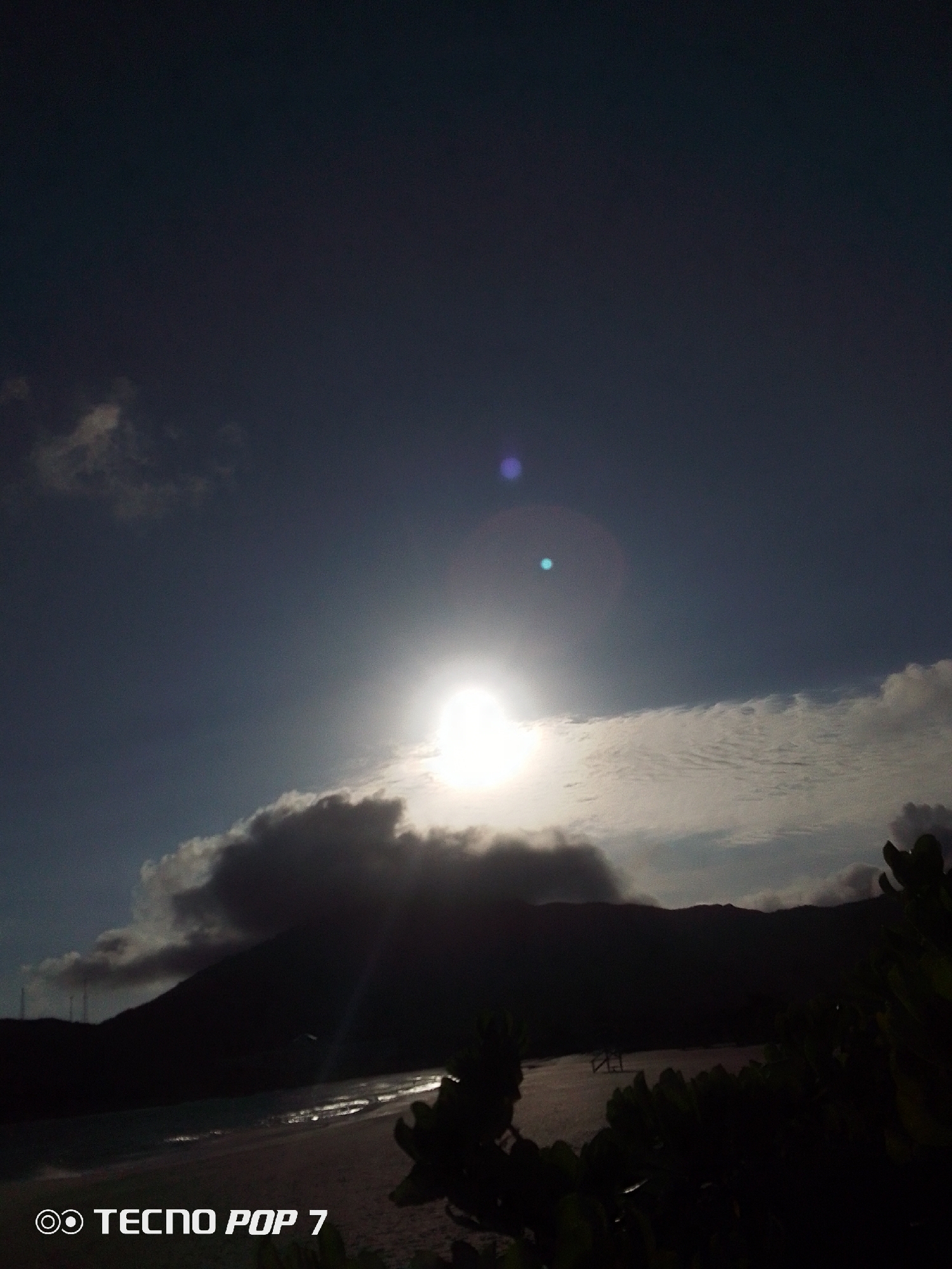 The image depicts a landscape in Pedro González, Venezuela, with a bright sun shining behind a layer of clouds. The sun's glare creates lens flare artifacts in the sky. The horizon is defined by dark, silhouetted mountains and what appears to be a coastline with a beach. Vegetation is visible in the foreground, obscuring some of the view. A structure, possibly a dock or pier, is partially visible in the water. The overall scene is dark due to the sun being the primary light source and silhouetting the foreground and distant features. The text "TECNOP POP 7" is visible in the bottom left corner, indicating the device used to capture the image.