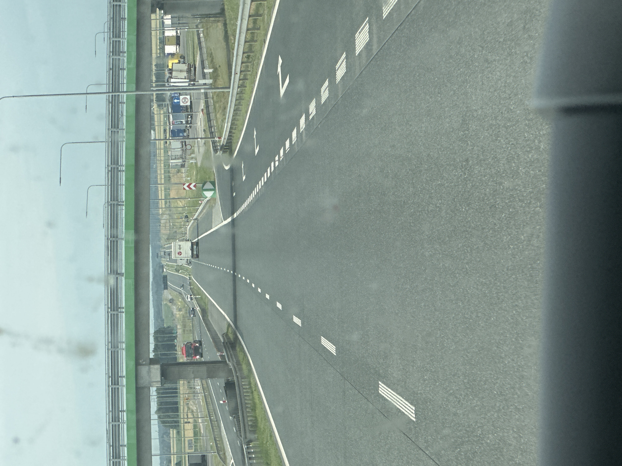 The image depicts a highway scene in Zabiele, Poland, with a focus on the road and its surroundings. The foreground shows the asphalt of the highway, marked with white dashed lines and solid white lines. In the middle ground, several vehicles are visible, including a red truck and a white camper van driving on the highway. An overpass, painted green and grey, spans across the highway in the background. On the right side of the highway, a rest stop or parking area is visible, with several trucks parked. The sky is overcast. The image appears to be taken from inside a vehicle, looking forward.