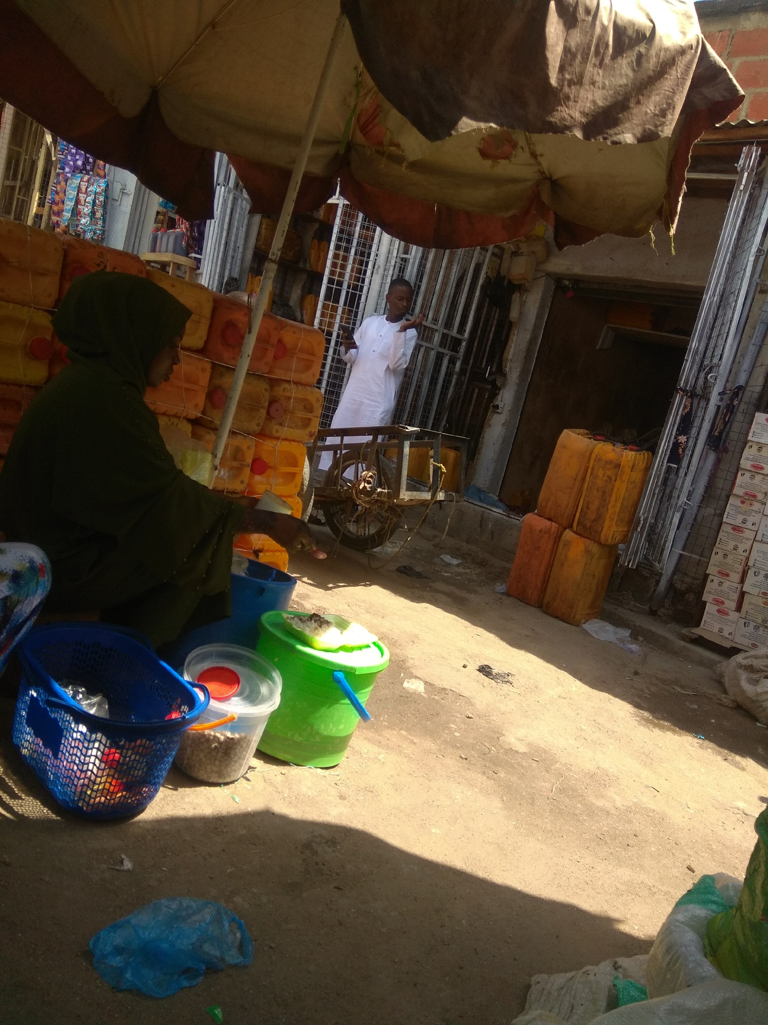 The image depicts an outdoor market scene in Jalingo, Nigeria, under a large, striped umbrella. In the foreground, a person wearing a dark green hijab and robe is crouched down, appearing to interact with items in buckets. To their left, a blue plastic basket contains various items, some with colorful packaging. Next to the basket are two translucent buckets, one with a red lid and the other filled with what appears to be small grains or beads, and a large green bucket with a white lid, containing some food items on top. A blue plastic bag lies on the sandy ground in front of these. In the background, a man in a white robe stands near a doorway, and stacks of large yellow and orange plastic containers are visible. Further back, rows of boxed goods are stacked along a wall. The ground is unpaved and sandy, with shadows cast by the umbrella and structures.