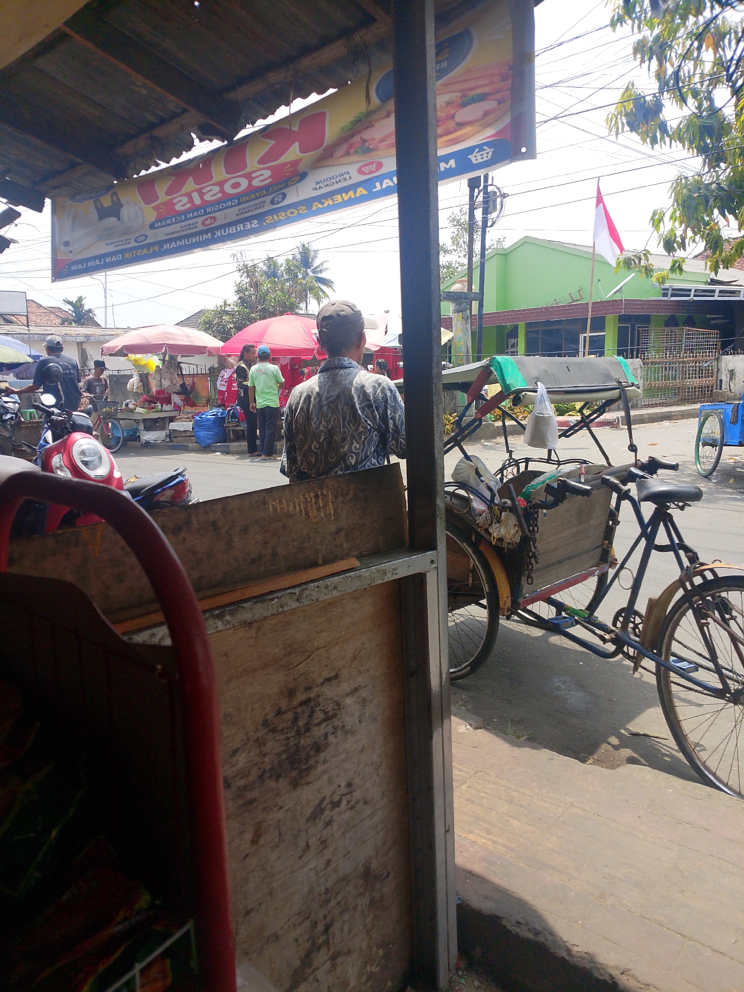 The image depicts a street scene in Palembang, Indonesia, on a sunny day. A man, seen from the back, is standing near a wooden structure, possibly a market stall. In the background, there are several market stalls with red and yellow umbrellas, suggesting a bustling marketplace. A traditional Indonesian three-wheeled bicycle, known as a becak, is parked on the side of the road. The becak is blue with a wooden cargo box and a canopy. Several motorcycles are also visible, indicating the presence of traffic. A green building with a red-tiled roof stands prominently in the background, and an Indonesian flag is flying in front of it. The overall environment appears to be a typical street in a commercial area, with elements of local culture and daily life.