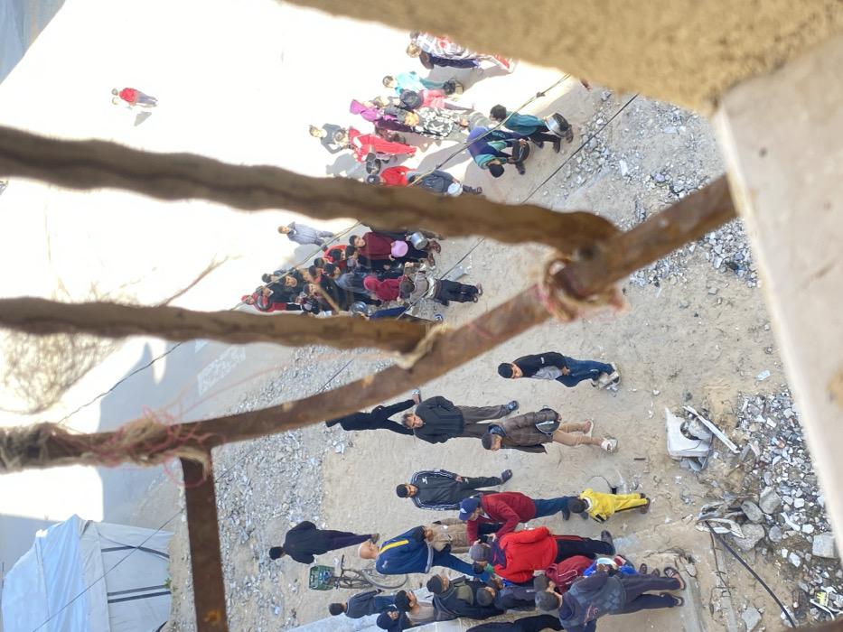 This elevated, downward-looking image captures an outdoor scene in Khan Yunis, Palestinian Territory. The immediate foreground is blurred by three vertical, gnarled brown elements, possibly rusted rebar, with pink twine wrapped around one. A light-colored concrete ledge is visible at the bottom right.
The scene below is divided into two primary areas. To the left, on unpaved sandy ground mixed with light-colored rubble, a smaller group of approximately 10-15 males, including men and boys, is gathered. One man wears a blue and yellow jacket. A person in dark clothing walks towards the center from the upper left, where a white tent structure with dark vertical stripes is partially visible. A bicycle with a green crate is parked nearby.
To the right, a larger gathering of approximately 30-40 individuals, predominantly children and adolescents, is arranged along a paved road or path. Many in this crowd are holding metallic or plastic bowls and containers, indicating a likely distribution event. Individuals are dressed in various casual attire, including jackets and hooded garments. The paved area continues into the bright background, showing a few distant figures. The central foreground features exposed dirt and scattered rubble, with visible utility lines or cables crossing the lower portion of the frame. Strong sunlight illuminates the scene, creating distinct shadows.