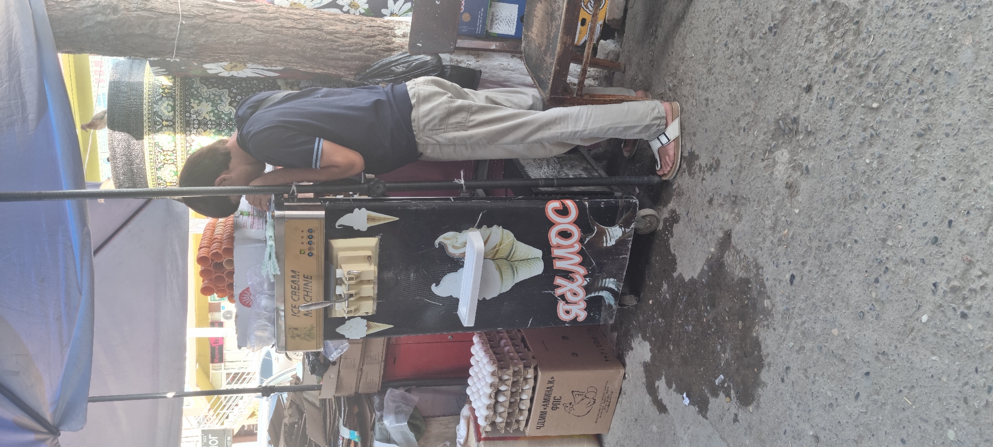 A young man wearing a dark blue t-shirt and light beige trousers stands at an outdoor ice cream stand in Bokhtar, Tajikistan. He is leaning over an ice cream machine, his face obscured. The machine is black and silver, with a dispenser on top and graphics of ice cream cones and the Cyrillic text "ЯХМОС" (Yakhmoc) on the front. To the left of the machine, there are stacks of egg cartons, one of which has text and a graphic of a chicken. In the background, an awning covers the stand, and behind it, buildings are visible. A tree with a floral pattern painted on it is to the right of the ice cream machine. The ground is a rough, grey concrete surface with some damp patches and debris. A pigeon is perched on a dark, textured surface in the upper background.