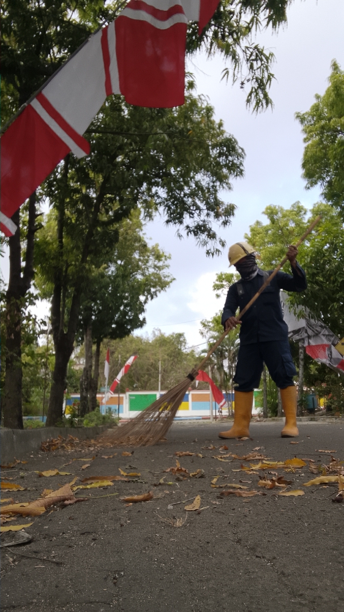 In Tlogobendung, Indonesia, a person wearing a dark blue uniform, a yellow hard hat, and bright yellow boots is sweeping a paved surface with a broom. The broom's bristles are made of dried grass or straw, and it has a long wooden handle. The person is also wearing a dark fabric mask that covers their lower face. Scattered on the ground are dry, fallen leaves. In the background, there are trees with green foliage and a clear sky. Several Indonesian flags, red and white, are visible, with one prominent flag attached to a pole in the upper left quadrant of the image and others in the background. Behind the sweeping area, there is a low wall or curb, and further back, a colorful structure with horizontal stripes of white, red, green, and blue. The overall scene appears to be an outdoor public area, possibly a park or roadside, during the daytime.