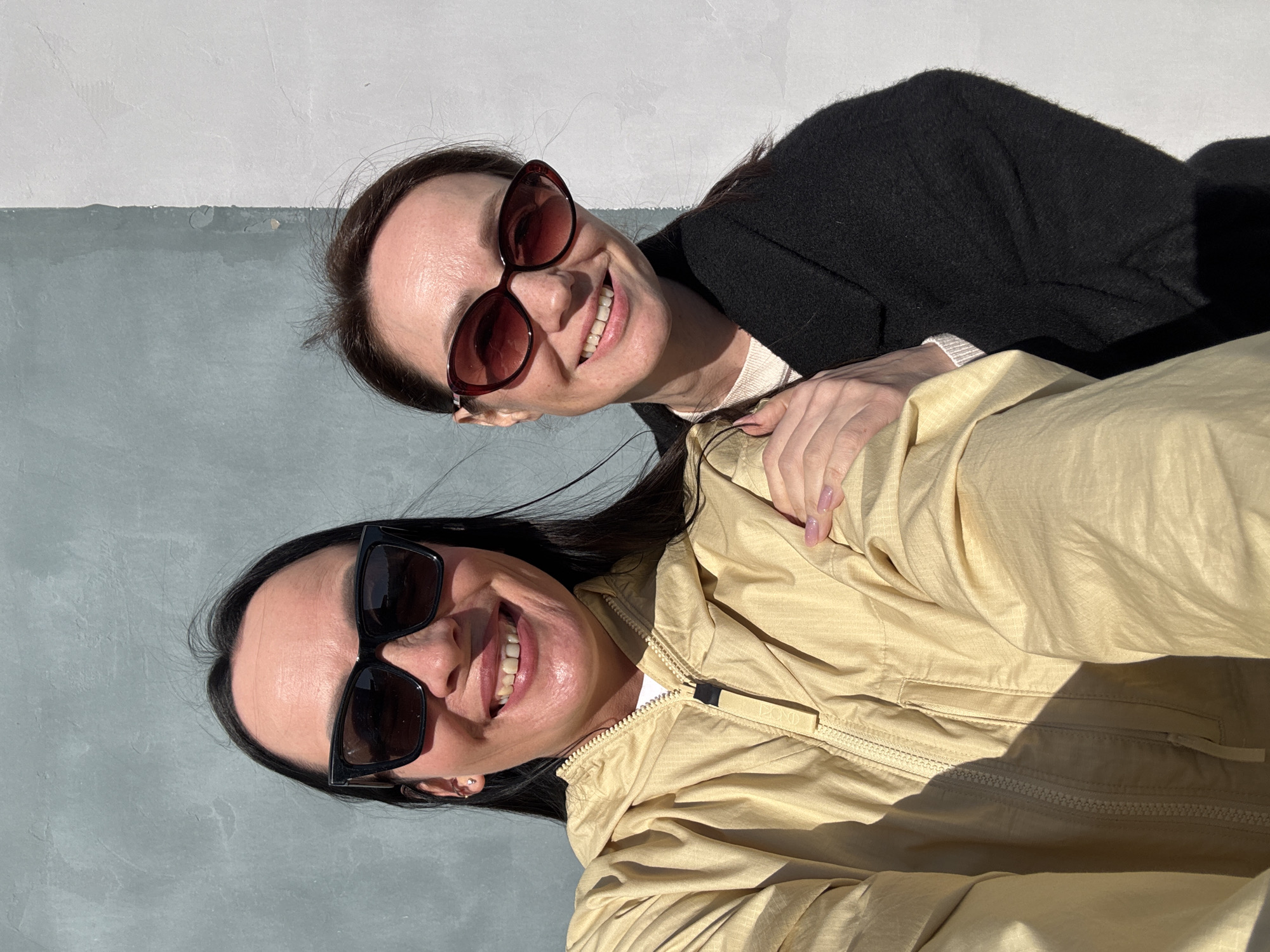 Two women are posing for a photo outdoors against a wall that is painted gray on the bottom and white on the top. The woman in the foreground has dark hair and is wearing a light tan jacket and dark sunglasses. She is smiling broadly. The woman in the background has brown hair and is wearing dark red sunglasses and a black sweater. Her hand is on the shoulder of the woman in front. Both women appear to be happy. The location is Chaikovsky, Russia.