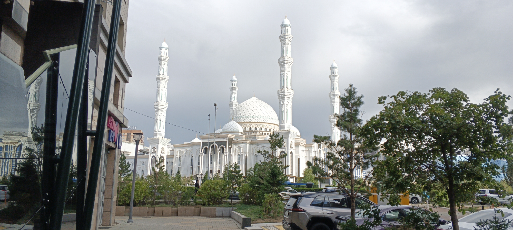 The image shows the Hazret Sultan Mosque in Astana, Kazakhstan, on a cloudy day. The mosque is a large, white building with multiple domes and minarets. It is surrounded by greenery and trees. In the foreground, there is a modern building with reflective windows that mirror the mosque. Several cars are parked on the street in front of the mosque. A lamppost is visible on the sidewalk. The overall scene suggests an urban environment with a prominent religious landmark.