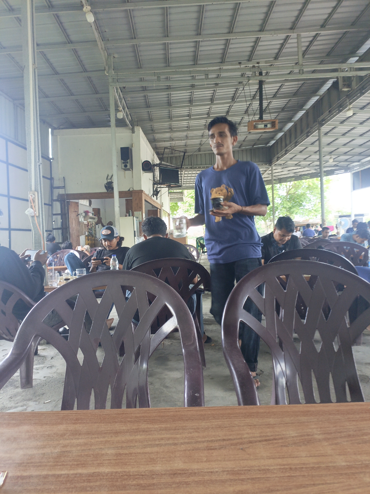 The image depicts an outdoor seating area of a cafe, likely in Lhokseumawe, Indonesia, with a corrugated metal roof supported by metal beams. In the foreground, two brown plastic chairs are visible, and beyond them, a wooden tabletop. A man in a blue t-shirt with a teddy bear graphic and dark pants is standing, holding a glass and a small item, possibly a toy. He appears to be presenting these items. Several other individuals are seated at tables in the background. One man wears a cap and is looking at his phone. Another man in a black t-shirt is also seated. The overall environment suggests a casual, open-air cafe setting.