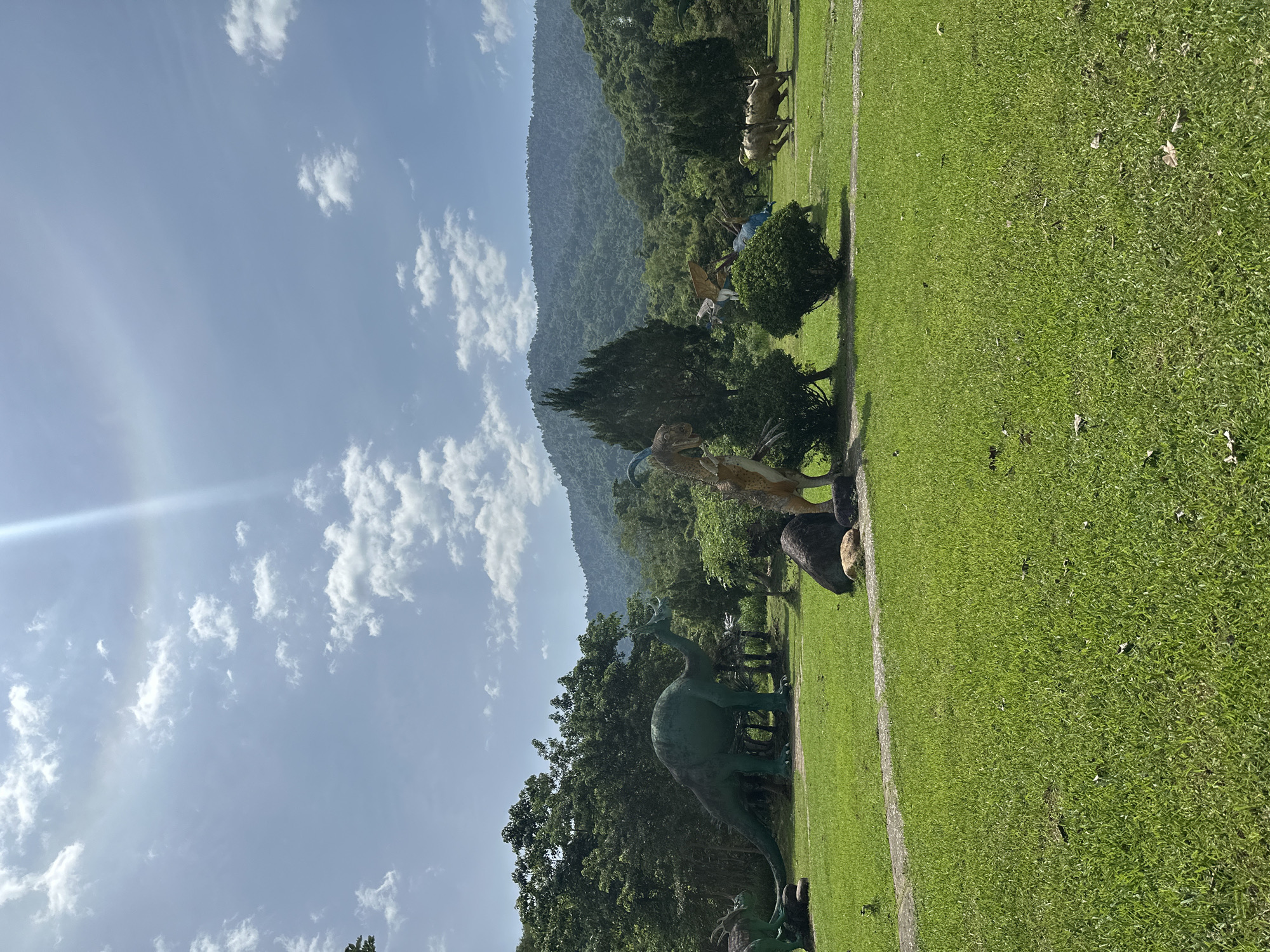 The image depicts a park-like setting with several dinosaur statues placed on a grassy field. In the foreground, a lush green lawn stretches across the frame, with a stone pathway winding through it. In the mid-ground, various dinosaur figures are scattered amongst trees and bushes. A large green brachiosaurus statue stands prominently on the left, while other dinosaur models, including what appears to be a velociraptor and a triceratops, are visible further back. The background is dominated by a dense forest that gives way to rolling hills and mountains under a bright, partly cloudy sky. The sun is visible, casting a lens flare across the upper portion of the image. The scene is characteristic of a tourist attraction or theme park, likely located in or near the city of Khanh Vinh, Vietnam, given the lush vegetation and mountainous landscape often found in the region. There are no people visible in the image.