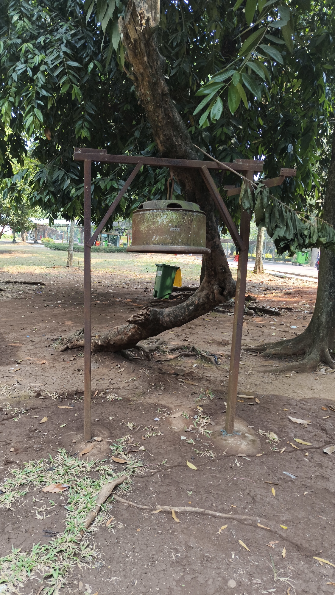 The image depicts an outdoor scene, likely a park or public space, in South Jakarta, Indonesia. A rustic metal stand holds a large, circular, weathered object, possibly a bell or a heavy decorative element, suspended above the ground. The stand is anchored into the soil with concrete bases. Behind the stand, a large tree with prominent roots grows, its trunk leaning at an angle. Two trash bins, one green and one yellow, are visible in the background. The ground is covered with dry soil, fallen leaves, and patches of sparse grass. In the far background, more trees and some structures can be observed, suggesting a populated area. The lighting indicates it is daytime, with shadows cast by the objects and foliage. No people are visible in the frame.