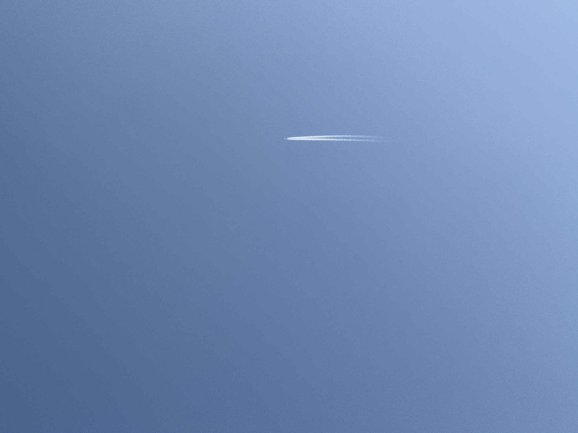 The image displays a clear, light blue sky over Izhevsk, Russia, with no ground features visible. A single, small white jet aircraft is present in the upper-middle section of the frame, depicted in flight. A distinct, linear white contrail trails vertically downwards from the aircraft, indicating its trajectory. No individuals or other objects are present in the scene.