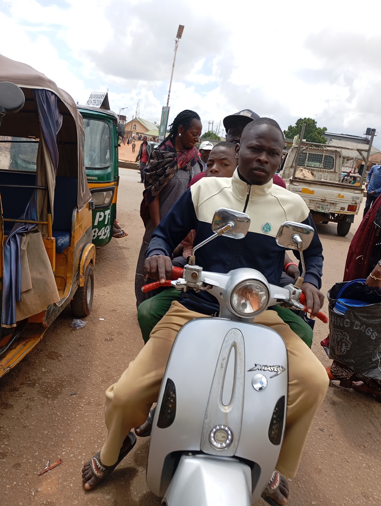 In Gura Top, Nigeria, a group of individuals are gathered on a dusty road. A man, wearing a light-colored jacket with dark blue accents and tan pants, sits on a silver scooter, looking directly at the camera. He has a serious expression. Behind him, another person, possibly a child, peeks over his shoulder. To the left of the scooter, a yellow and green auto-rickshaw, commonly known as an "okada" or "kabukabu" in Nigeria, is parked. The auto-rickshaw has blue seating and a brown canopy. In the background, several other people are visible, some walking and others standing near buildings and a white truck. The sky is partly cloudy, suggesting it might be a warm day. One of the people in the image is wearing a dark patterned scarf, and another person is wearing a blue shirt. A black plastic bag with "GOOD MORNING BAGS" written on it is visible on the right side of the frame. The overall scene depicts a bustling street in a Nigerian town.