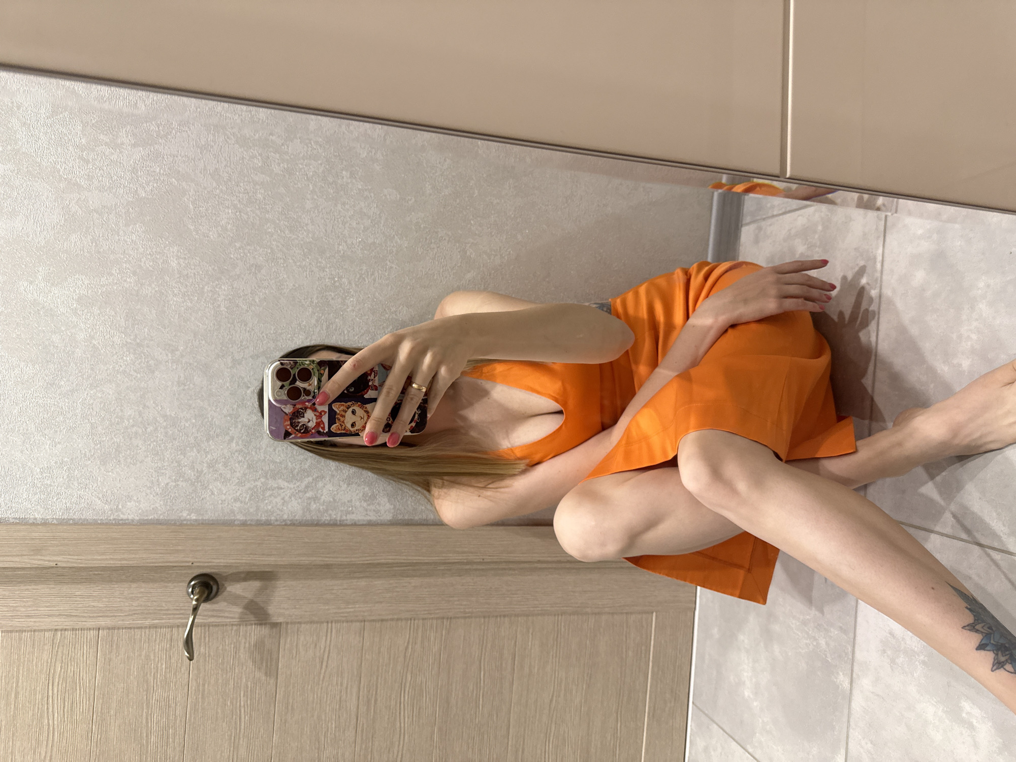 The image shows a woman in an orange dress taking a selfie in a bathroom. She is sitting on the floor, with her legs crossed. The woman's phone is visible, and it has a case with cat designs. Her nails are painted pink. A tattoo is visible on her leg. The bathroom has tiled walls and a wooden door with a silver handle. The lighting suggests it is daytime, and there are no indications of the weather outside. The scene appears to be in a domestic setting.