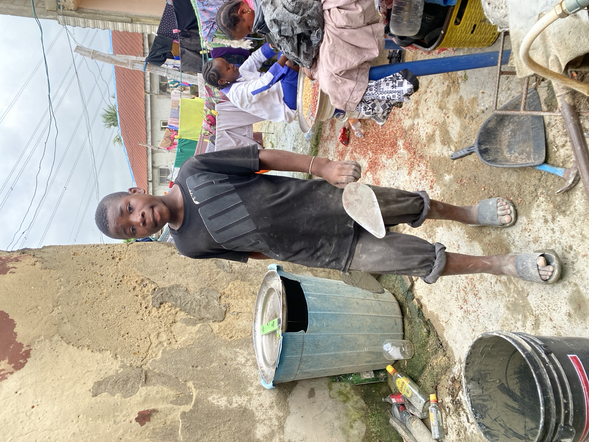 A young boy stands in a yard with a textured, painted wall behind him. He holds a trowel. Two young girls are sitting and looking at a large metal bowl of peanuts. Spilled peanuts are scattered on the ground. Clothes are hanging on a line in the background. Various containers are near the boy. Ado, Nigeria.