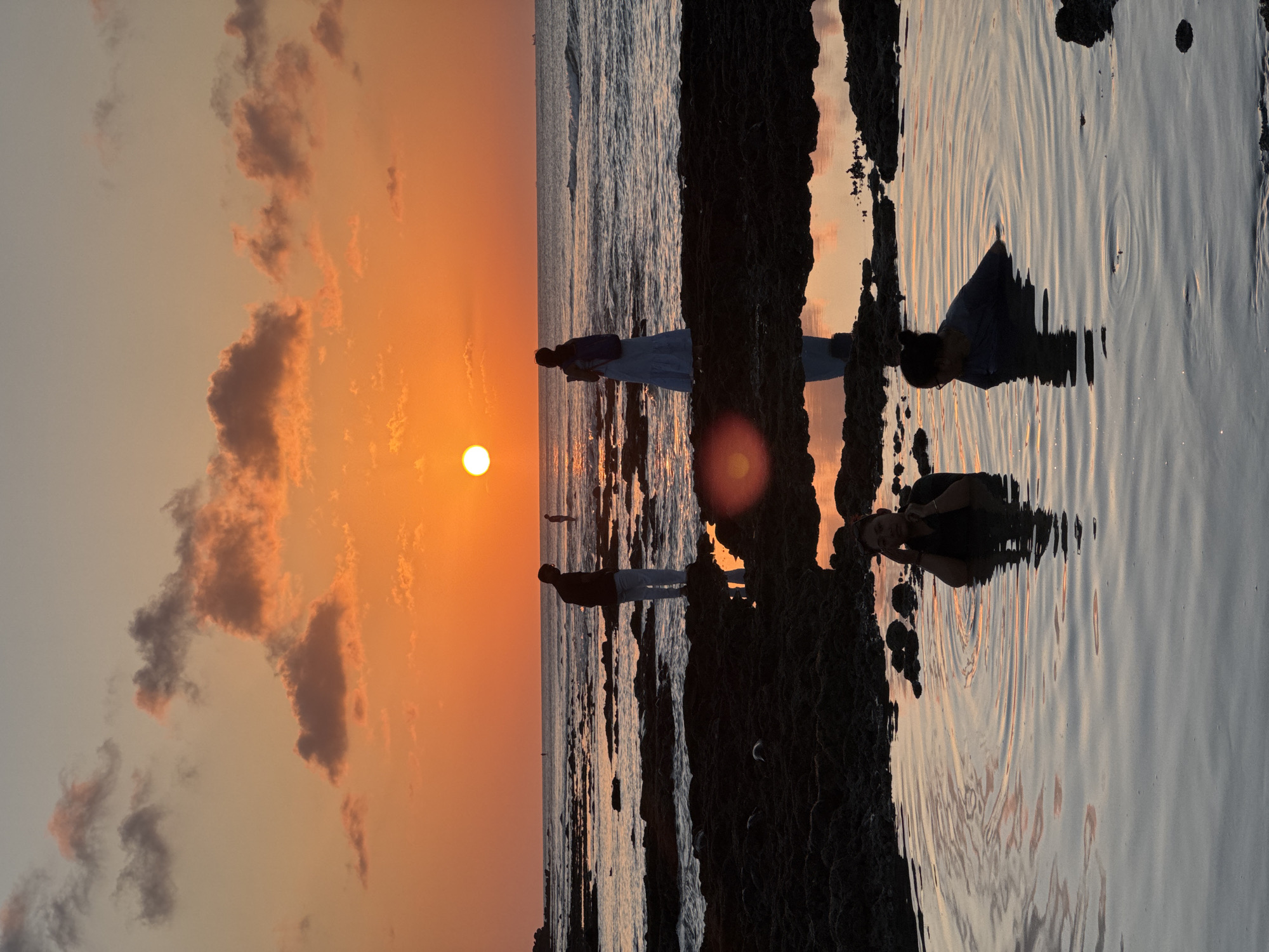 The image depicts a serene sunset scene at the rocky shoreline of Kuta Selatan, Indonesia. The sun, a brilliant orange orb, hangs low in the sky, casting a warm glow on the scattered clouds and the rippling ocean waves. In the foreground, two individuals are partially submerged in a calm tidal pool, their bodies silhouetted against the vibrant sky. One person is seated, looking towards the camera, while the other is angled away, appearing to relax. Further back, on the rocky outcrops, two more figures stand near the water's edge. One, a man in light-colored pants, faces away from the viewer. The other, a woman in a light-colored dress and a backpack, stands in profile, looking out towards the sea. The rocky terrain is dark and wet, reflecting the colors of the sunset. The overall atmosphere is one of tranquility and natural beauty, capturing a moment of peaceful enjoyment of the coastal landscape.