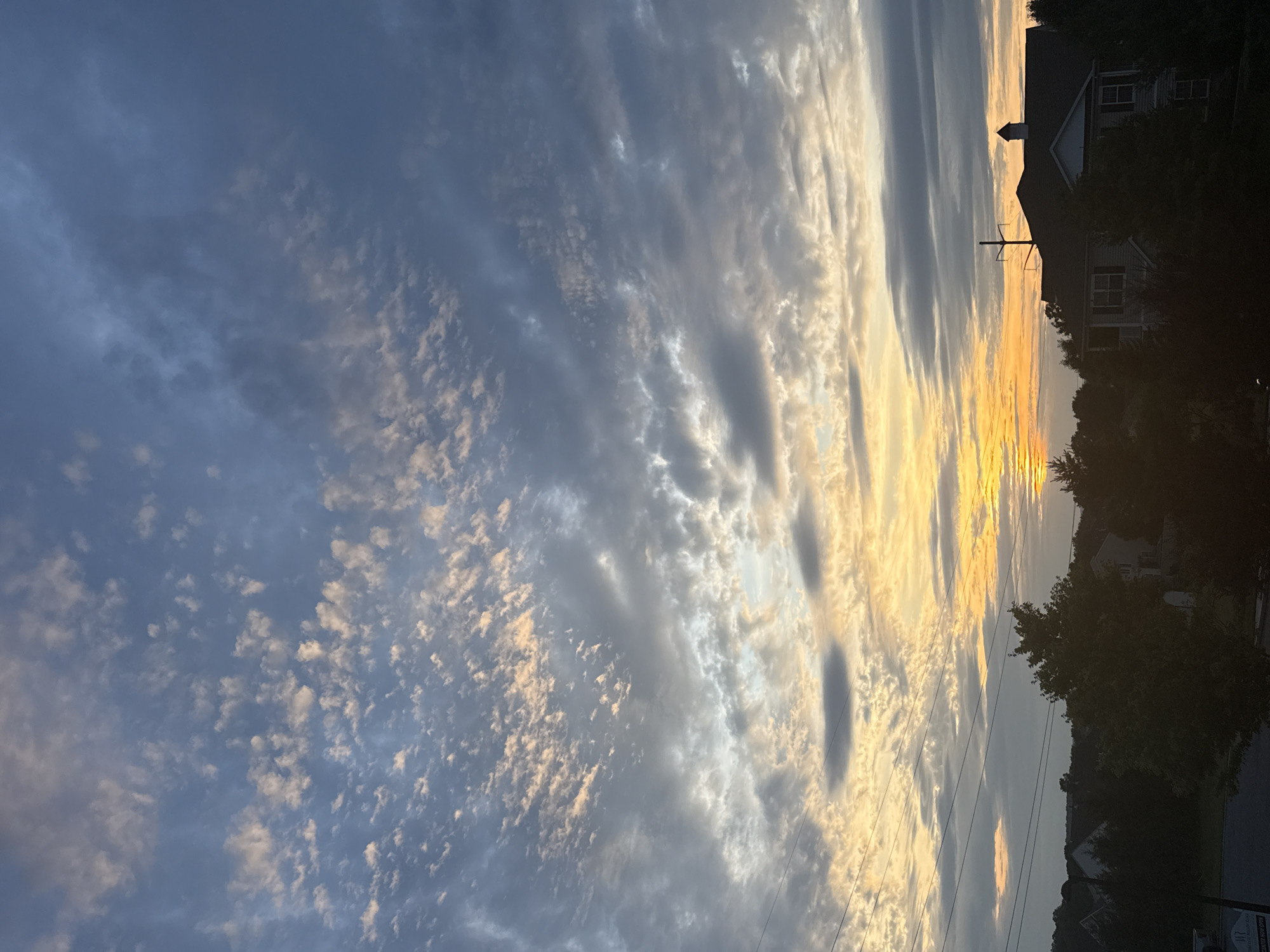 A dawn sky glows with soft oranges and yellows near the horizon, transitioning to a muted blue overhead. The sky is populated with wispy, scattered clouds, interspersed with darker, more solid cloud formations. Silhouetted against the vibrant sky are the dark shapes of trees and the rooflines of houses. One house, partially visible in the lower right quadrant, has a distinctive peaked roof and is surrounded by trees. Utility lines are visible stretching across the frame. The scene depicts a suburban landscape at sunrise in Allen Township, United States of America.