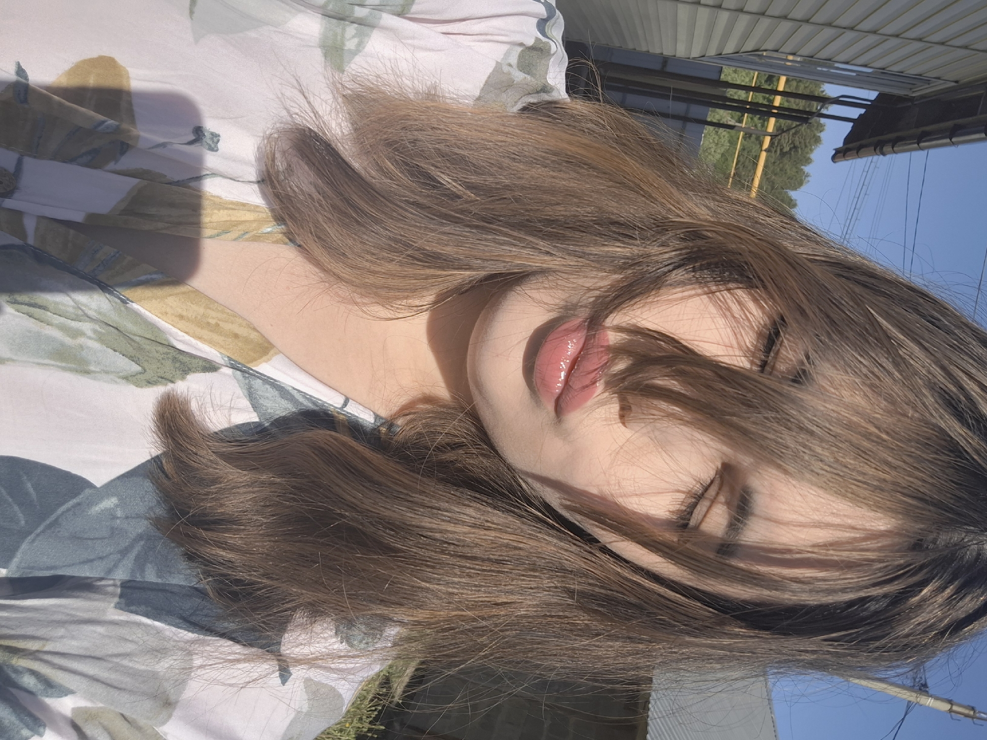 A person with long, light brown hair is lying down. The individual's eyes are closed, and strands of hair partially cover the face. The person has glossy lips and fair skin. They are lying on a floral-patterned fabric. The background shows an open area with metal structures, trees, and a blue sky. The location is likely outside under natural light. No information indicates this location is in the city of Амзя, Russia.