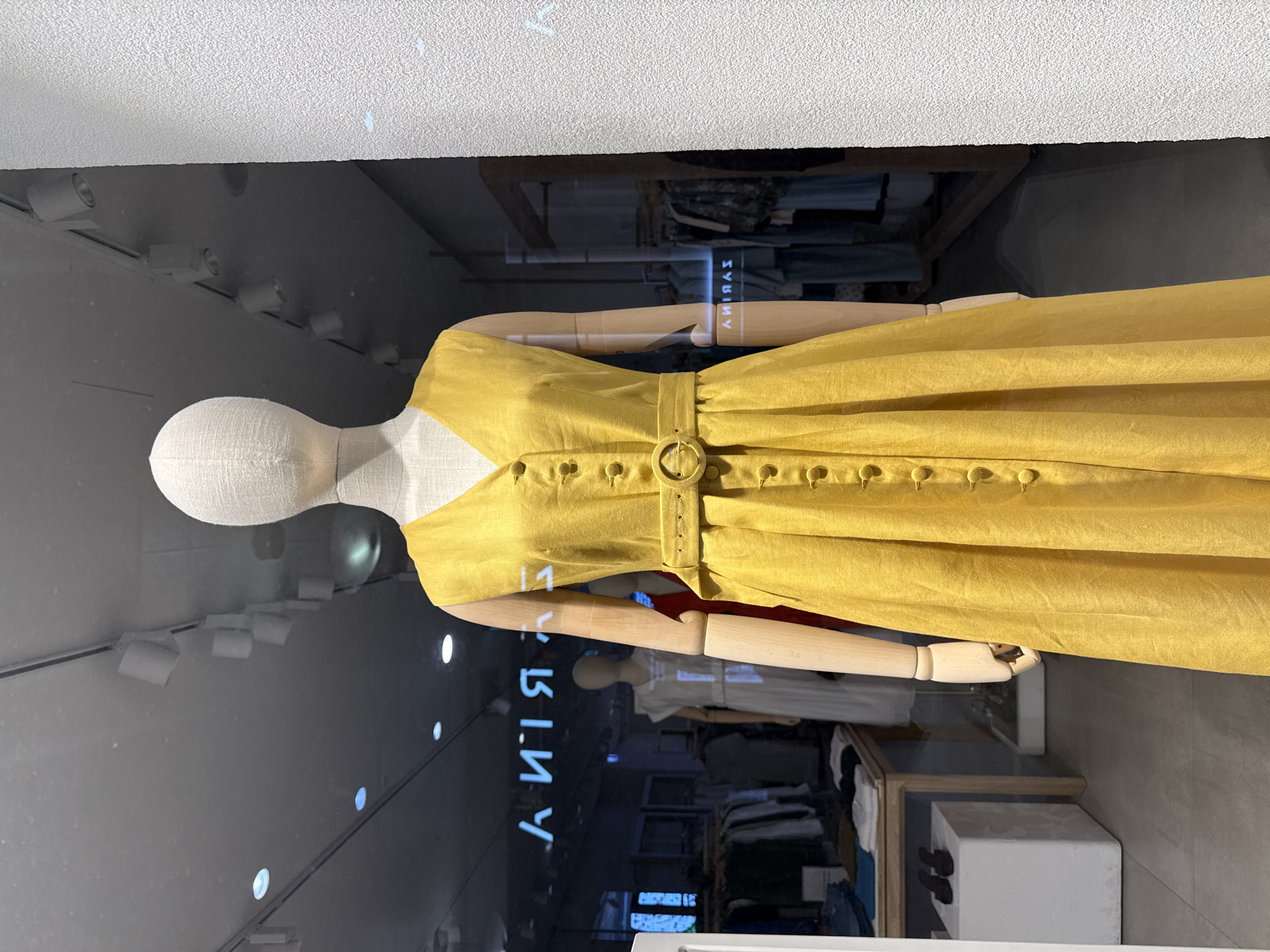 This is a medium shot, eye-level, vertical, indoor photograph of a mannequin in a store window display. The mannequin is dressed in a yellow, sleeveless, V-neck dress with a belted waist and button detailing down the front. The dress is made of a linen-like fabric and has a full skirt. The mannequin has a white, fabric-covered head and wooden arms. The store interior is visible through the window, with clothing racks and other mannequins in the background. The reflection of a blue neon sign with Cyrillic lettering is visible in the window, reading "А ИЛЯЯЯ". The background also shows a white textured wall on the right side of the image. The scene depicts a retail environment, likely a clothing boutique. The location is specified as Ukromne, Ukraine.