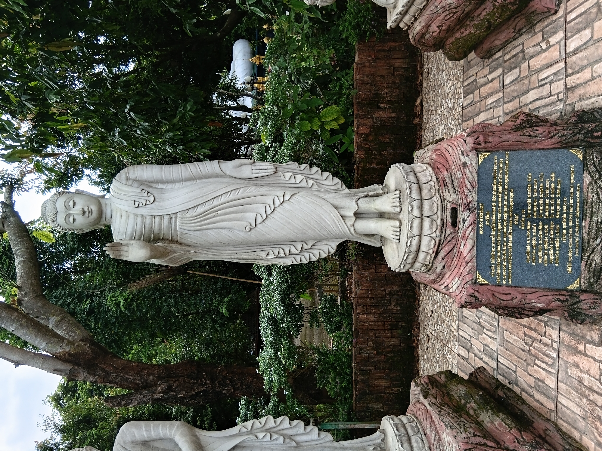 The image shows a statue of a standing Buddha in a garden setting at Ban Ruammit, Thailand. The Buddha statue is made of white stone and is depicted with its right hand raised in a gesture of blessing or protection. It is positioned on a decorative stone base. To the left and right of the main statue, parts of other stone statues are visible, suggesting a collection of statues. The background features lush green trees and foliage, indicating a natural, outdoor environment. A plaque with text, likely describing the statue or its significance, is situated at the base of the main statue. The ground is paved with patterned stones.