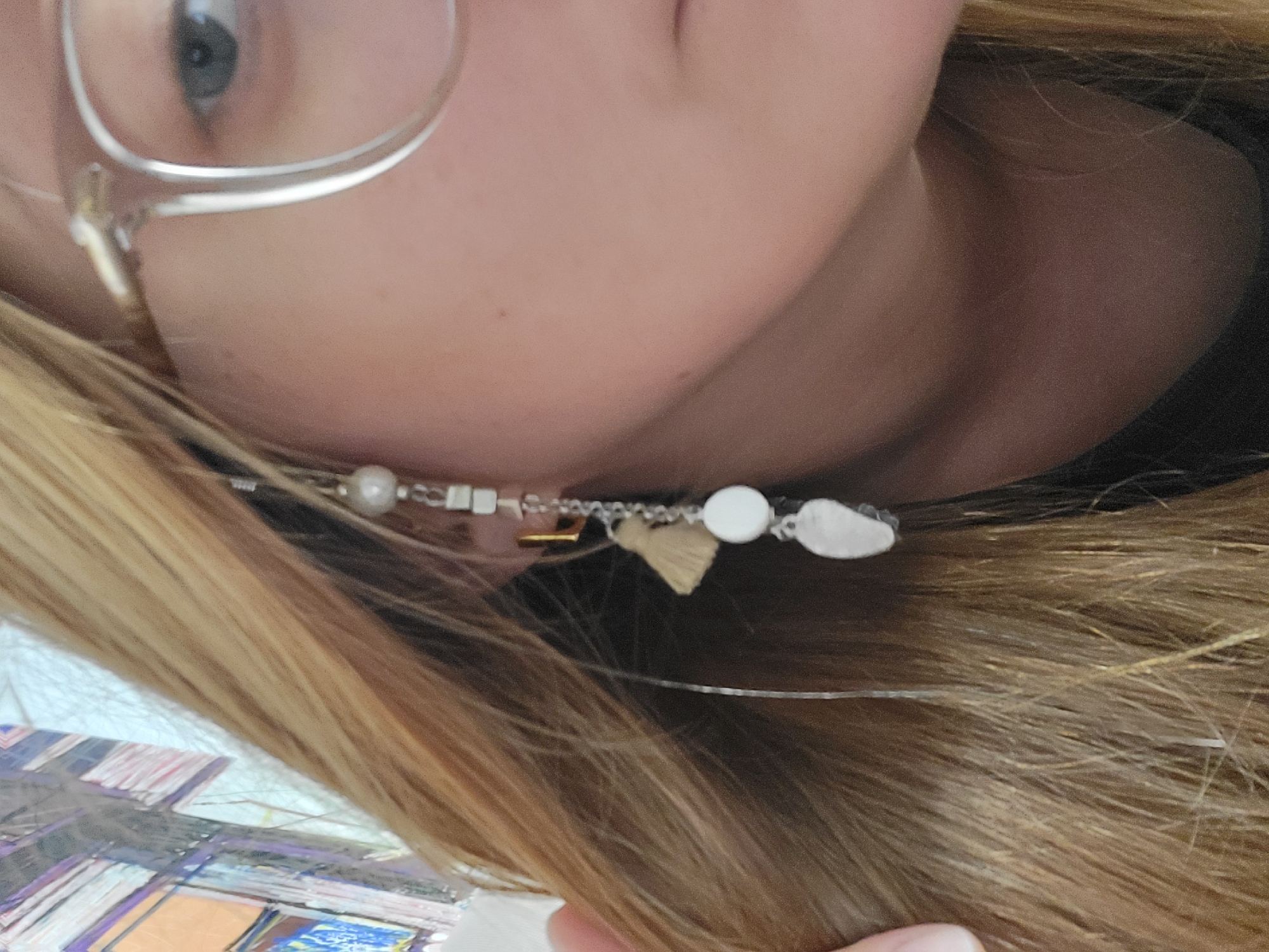 The image shows a close-up of a person's face, turned slightly to the side. The individual has fair skin, blonde hair, and is wearing glasses. A decorative hair accessory with beads and charms is visible in their hair. The setting is unclear, and there is no information to suggest a location in Budva, Montenegro.