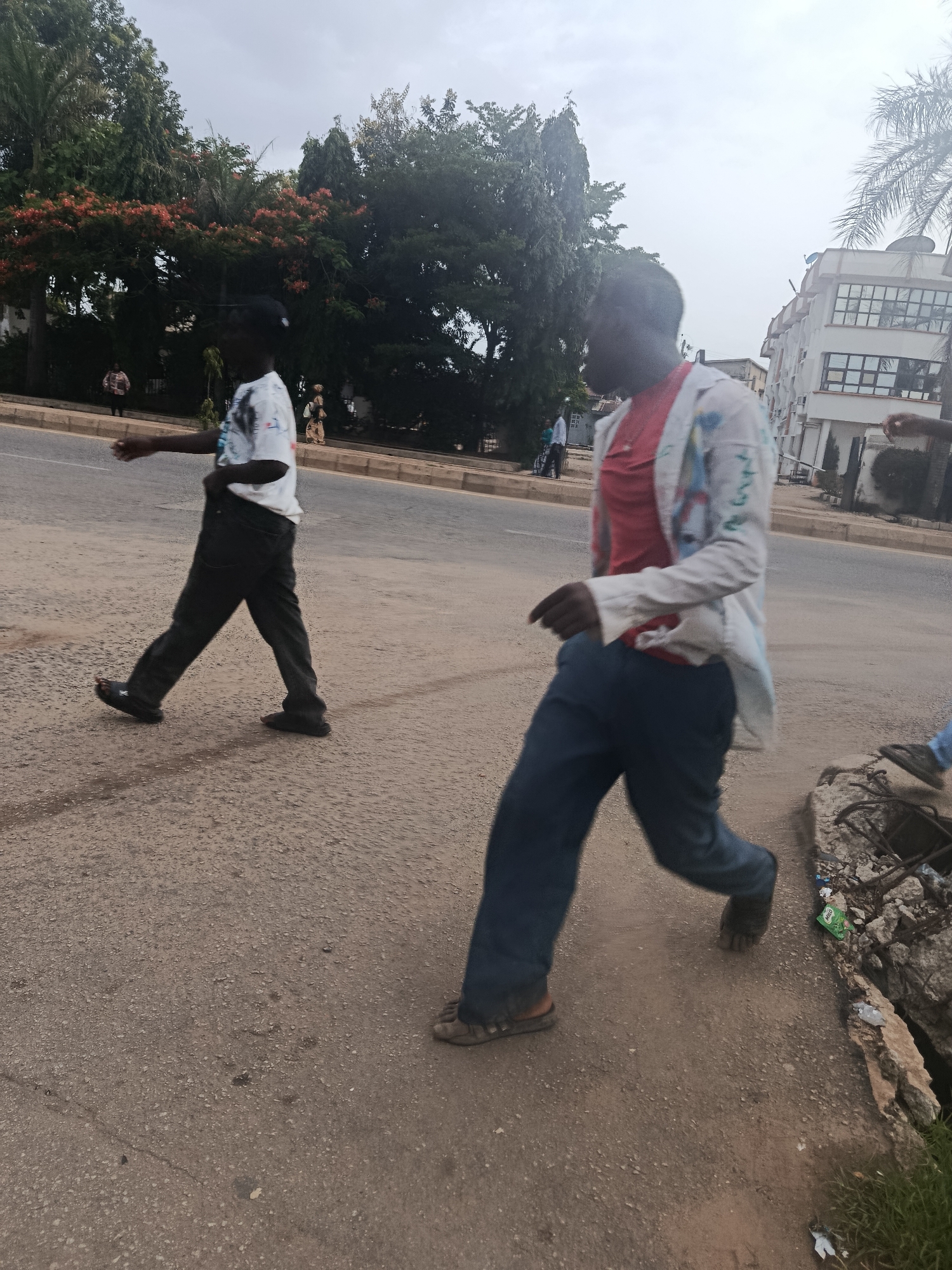 Two individuals walk on a paved street in a city environment, possibly New Stadium, Nigeria. The person in the foreground, on the right, wears a white jacket with colorful designs over a red shirt and blue jeans. They are walking towards the right of the frame. To their left, the second person, wearing a white t-shirt with graphics and dark pants, is walking in the same direction. In the background, there are trees with red flowers, a modern building with multiple windows, and a palm tree. The street appears to be well-trafficked, with a broken curb and some debris on the right side of the road. The sky is overcast.