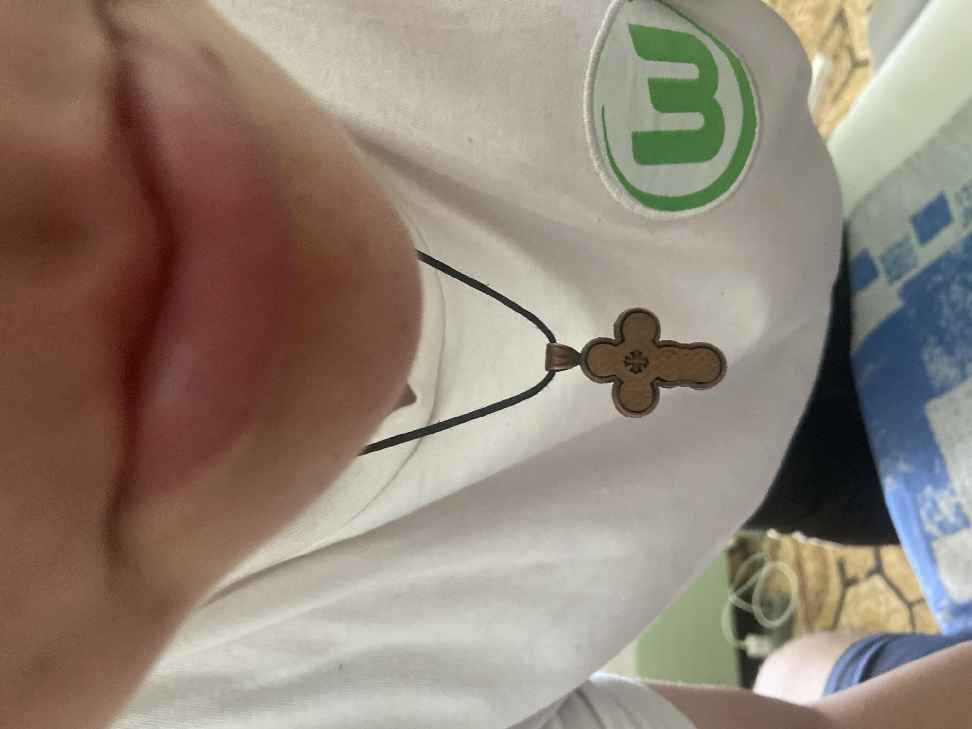 The image shows a close-up view of a person wearing a white t-shirt with a necklace and a patch. The person's lips are visible in the upper part of the frame. The necklace has a wooden cross pendant. On the right side of the t-shirt, there is a circular green patch with a white stylized 'W' inside. The background is blurred and appears to be an indoor setting with some electrical cords and patterned flooring. The image quality is moderate, with some noise and a slight blur. The scene is static and does not depict any specific action or interaction.