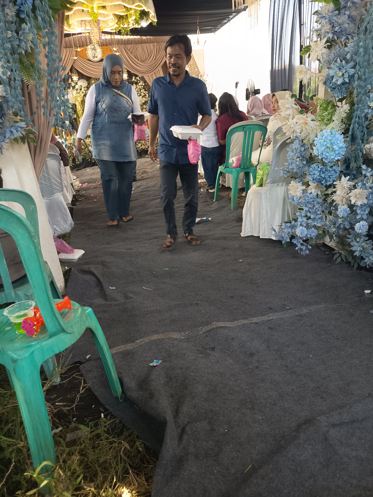 In an outdoor event space in Unknown, Unknown, a middle-aged man wearing a blue button-down shirt and dark pants walks across a dark carpeted aisle. He carries a white food container in his right hand and a pink plastic bag in his left. To his left, a woman wearing a blue denim vest over a white long-sleeved shirt and a grey hijab also walks, holding a white food container in her right hand and a pink plastic bag in her left. She is wearing blue jeans and dark sandals. Behind them, several guests are seated at tables draped in white cloths, some with blue and white floral arrangements. A bright green plastic chair is visible in the foreground, with a clear plastic cup containing a green liquid and a toy on it. The aisle is lined with chairs and draped fabric, creating a festive atmosphere. The ground on the left side of the aisle is covered with grass.