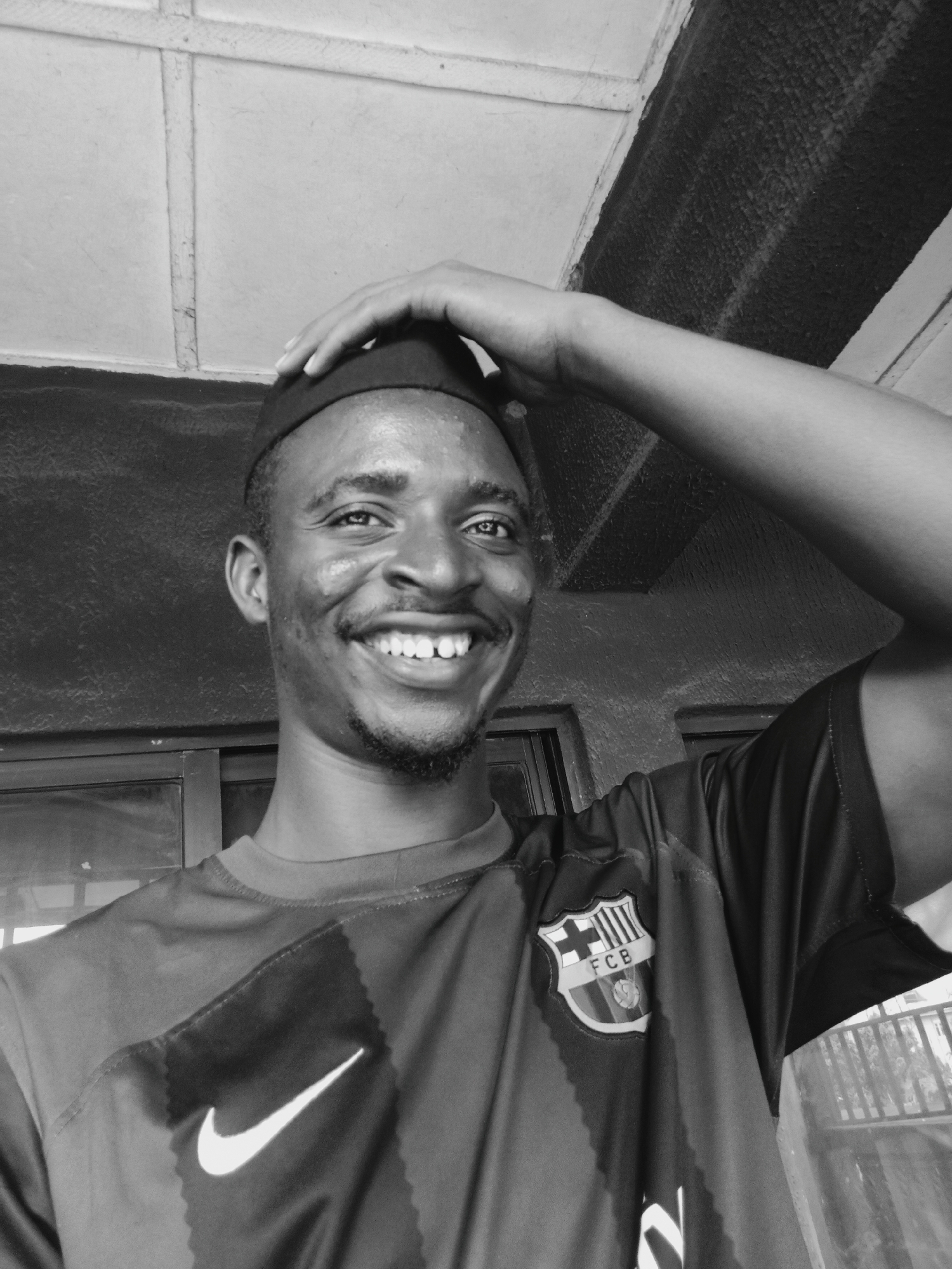This black and white image captures a smiling young man in a close-up portrait. The subject, likely in Onitsha, Nigeria, wears a dark, close-fitting cap on his head, with his right hand gently resting on it. He exhibits a broad, genuine smile, revealing his teeth, and his eyes also convey a cheerful emotion. A short beard and mustache frame his face.
He is dressed in a short-sleeved football jersey, predominantly dark in color. On the viewer's left side of his chest, a prominent white Nike "swoosh" logo is visible. On the viewer's right, the FC Barcelona crest is displayed, clearly showing the letters "FCB" within its design.
The background consists of a light-toned wall and ceiling structure with visible beams, indicating an indoor or sheltered outdoor setting, possibly a veranda or porch. Darker rectangular shapes, resembling windows or doors, can be seen in the background behind him. The lighting suggests it is daytime, and the overall mood of the portrait is one of happiness and approachability.