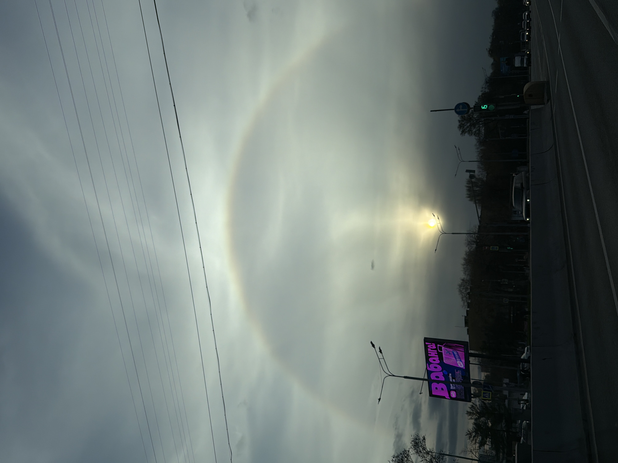The image shows a street scene in Moscow, Russia, on an overcast day. A distinct halo, a luminous ring, is visible around the sun. The halo occupies a large portion of the sky. Power lines stretch across the left side of the frame. On the right side, a road is visible, with cars, street lights, and traffic signals. A large electronic billboard with the word "ВВованга!" (V Vovanga!) written in a purple color can be seen on the roadside. Trees are visible in the background.