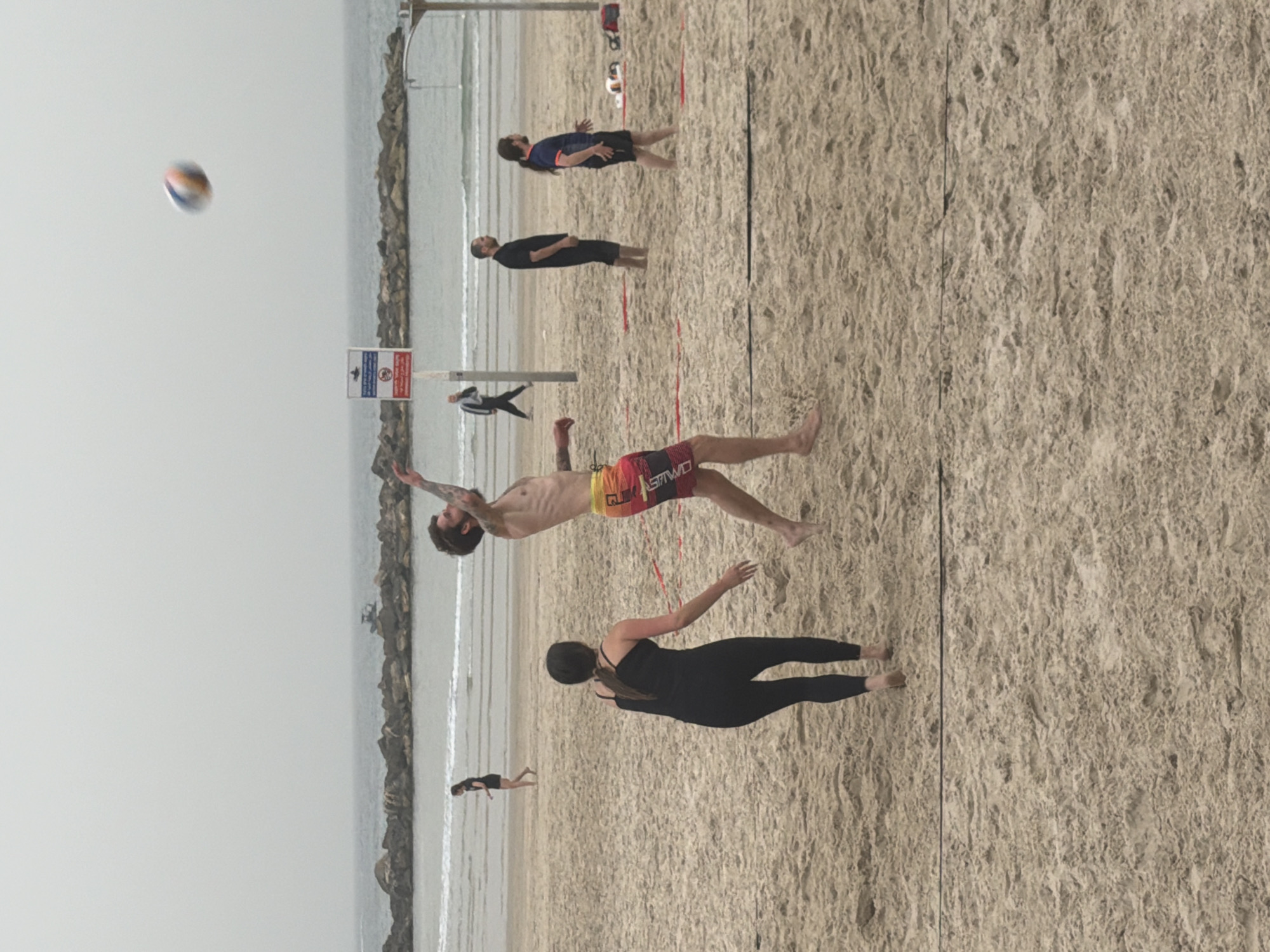 The image depicts a beach volleyball game in Tel-Aviv, Israel. A man with tattoos is jumping to hit the volleyball. A woman in black leggings and a top is preparing to receive the serve. Two other people are standing back further in the court watching, two more are further off to the side. The beach is sandy, with a volleyball net dividing the court. In the background, there are rocks along the shoreline and the Mediterranean Sea. The sky is overcast.