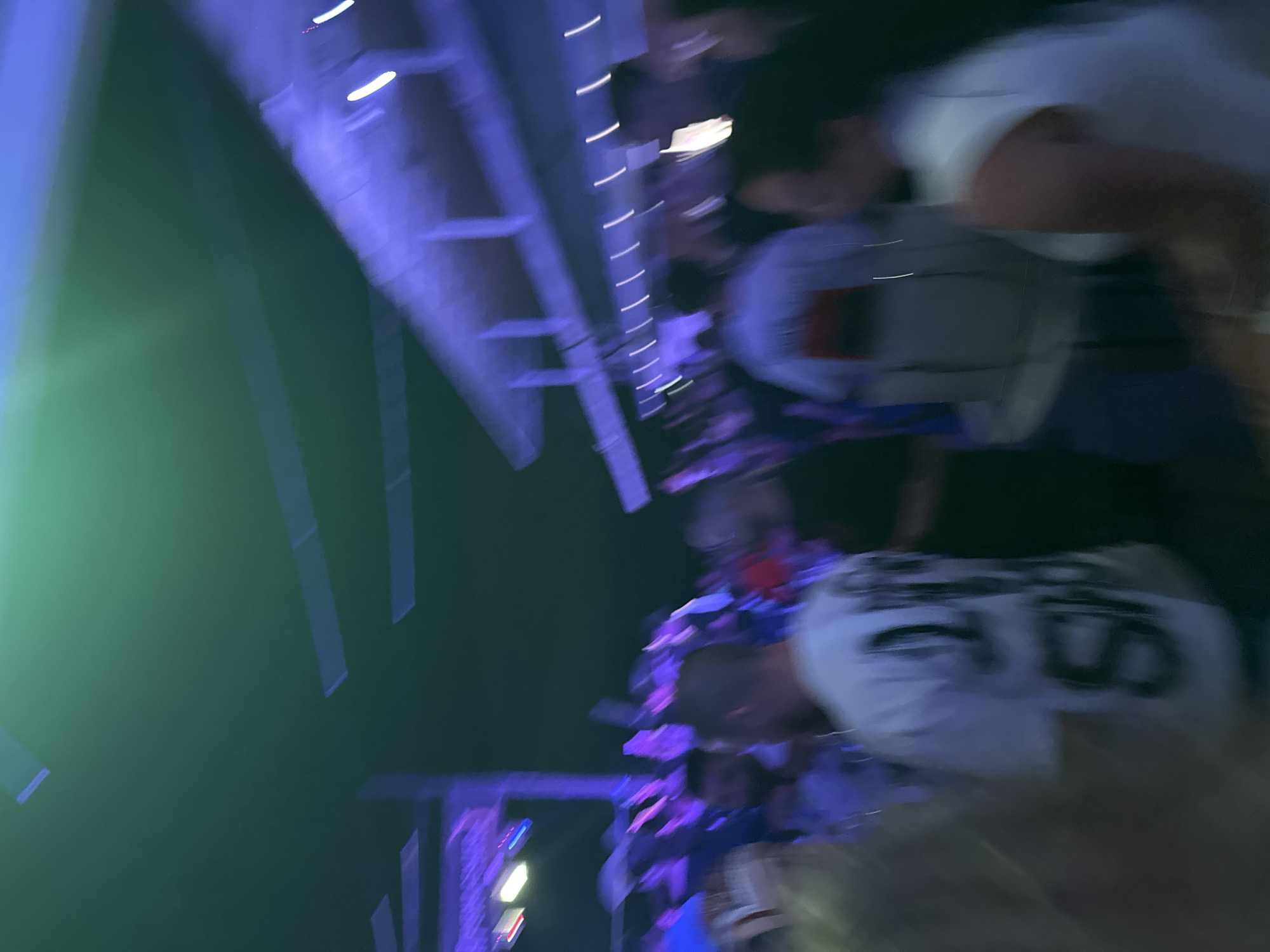 The image captures a blurred, nighttime scene of a crowded outdoor venue, possibly a concert or club, with vibrant colored lighting. A green light emanates from the upper left, casting a glow on the ceiling structure, which is illuminated by purple and white lights. Several people are visible, mostly out of focus, suggesting movement and a lively atmosphere. One individual in the foreground is wearing a white t-shirt with a black graphic design. The environment appears to be an open-air space with a covered section, suggesting it could be a festival ground or an outdoor entertainment complex. The context of Varna, Bulgaria, implies this scene is taking place within the city's entertainment district, likely during an event.