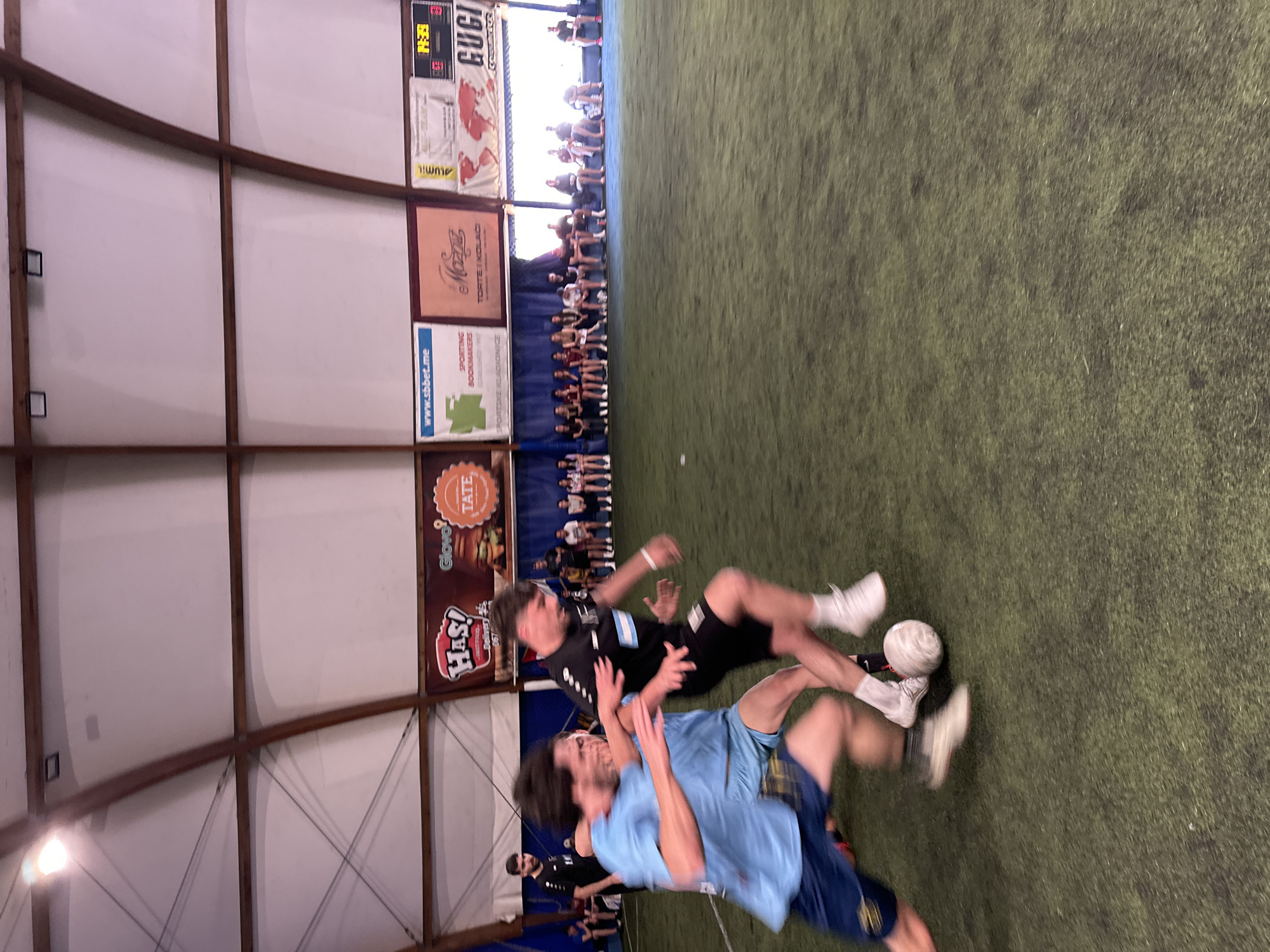 The image captures a moment during what appears to be an indoor soccer game. Two individuals are prominently featured mid-air, seemingly in contention for the ball. One person is wearing a black jersey and black shorts with white shoes, while the other is in a light blue shirt and dark blue shorts, also with white shoes. The ball is positioned between them, indicating an active play.
In the background, along the wall, there is a row of spectators seated, watching the game. The indoor setting suggests an arena or gymnasium with artificial turf as the playing surface. Various advertisements and posters are displayed on the walls, including brands like "HAS!", "Glovo", "TATE", "www.sbbet.me", and "Mozga." A scoreboard is also visible, displaying the time.
Based on the presence of the "www.sbbet.me" and "Mozga" advertisements, which are associated with Montenegro, and the context of indoor soccer, it is plausible that this scene is located in Budva, Montenegro.