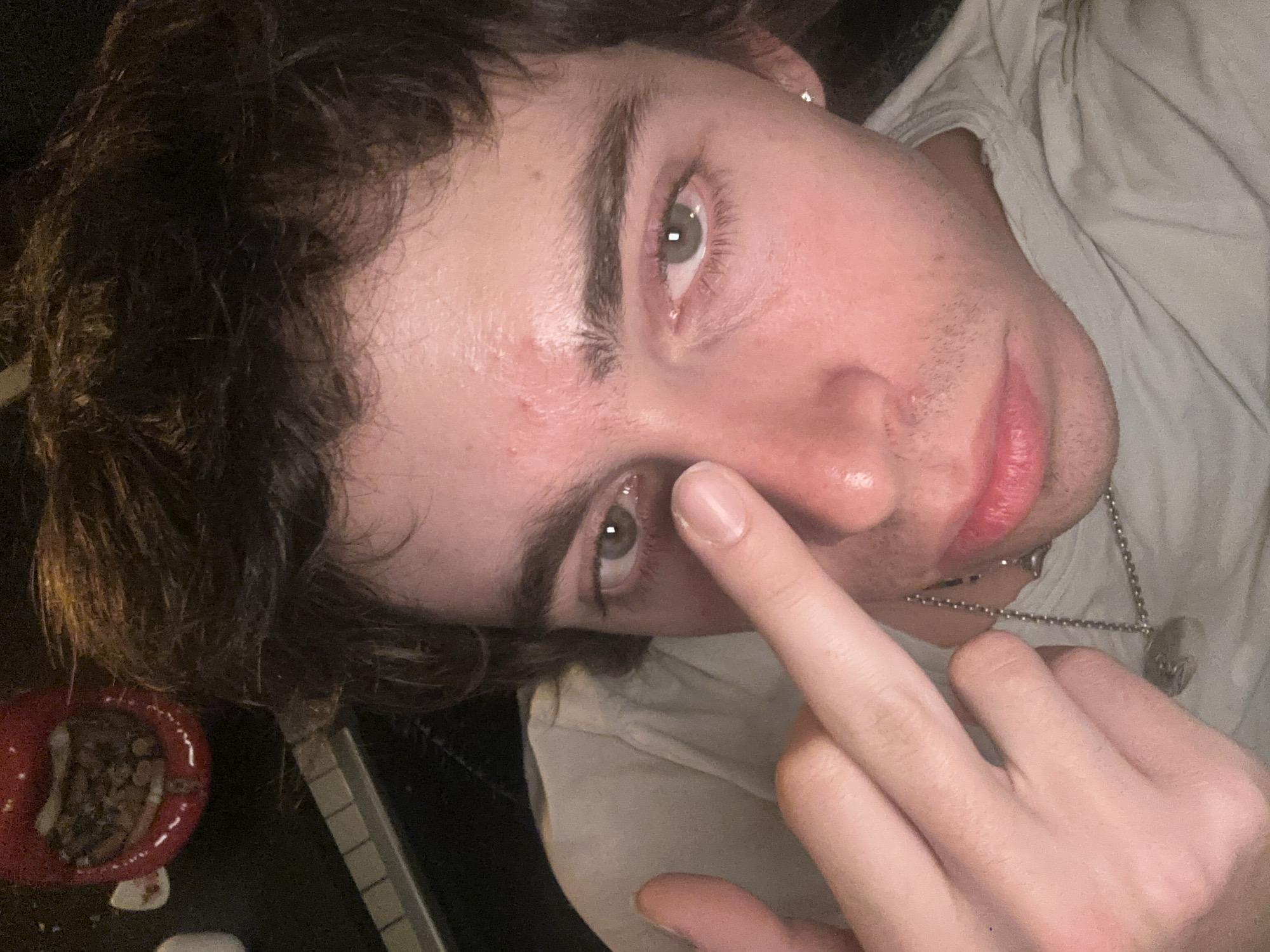 The image displays a high-angle, close-up selfie of a young male subject in an indoor, dimly lit setting. The subject, appearing to be in his late teens or early twenties, has messy dark brown hair, light green/hazel eyes, and fair skin with visible redness on his forehead and nose, alongside faint stubble on his upper lip and chin. He wears a light-colored t-shirt and a silver ball-chain necklace with a circular pendant. A silver stud earring is visible in his right ear. His left hand is raised, with the index finger touching the bridge of his nose, while the remaining fingers are curled. The subject gazes directly into the camera with a neutral expression. In the upper left background, a red, lips-shaped dish containing cigarette butts and ash is partially visible. Below this object, a dark surface with elements resembling the keys of a piano or synthesizer is discernible. The specific location within the city of Paris, France, cannot be determined from the visual evidence presented in the image.