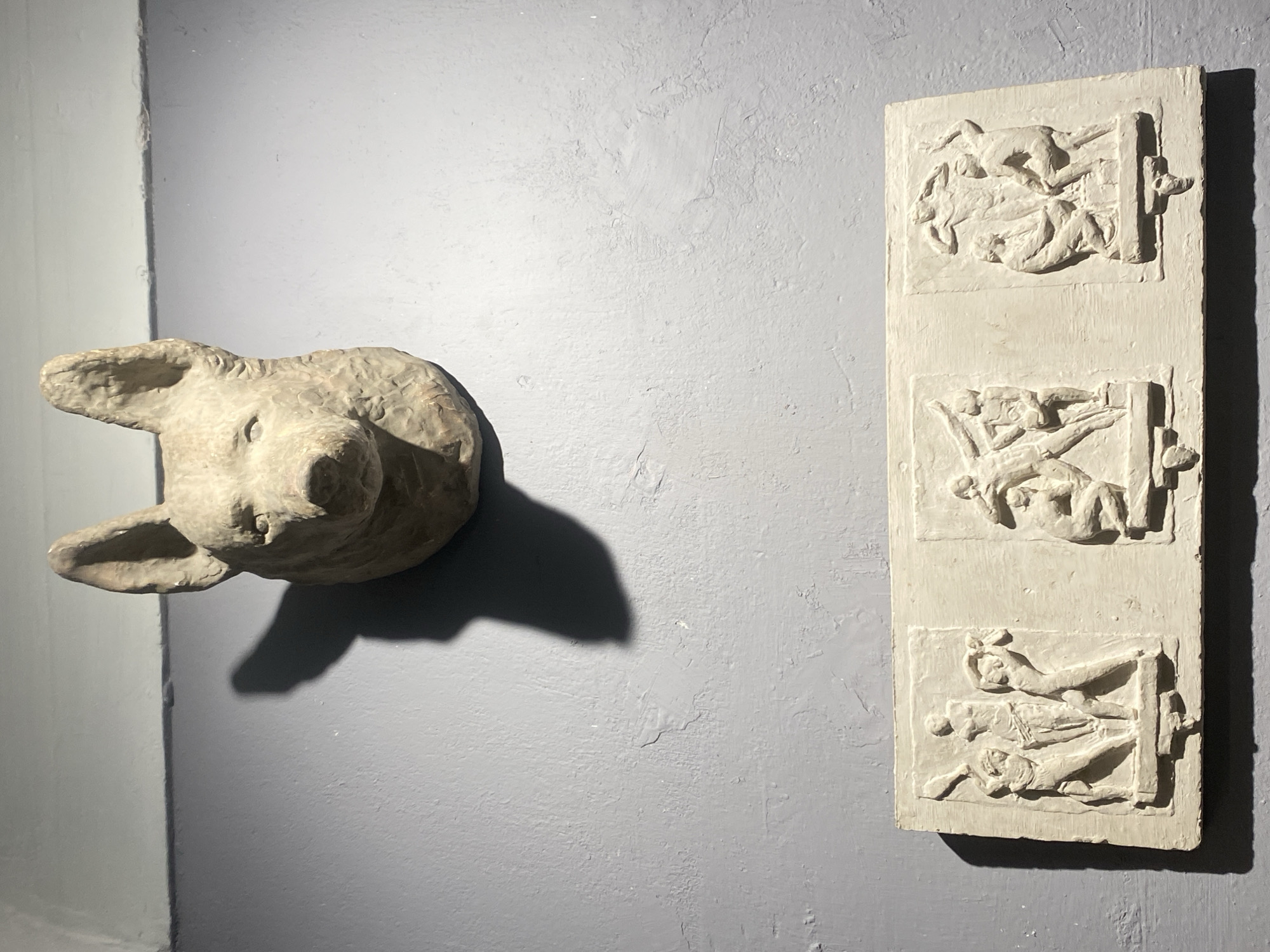 This image shows a light gray wall with two distinct art pieces displayed on it. At the top, a roughly textured, light-colored sculpture of a dog's head is mounted. It has prominent ears, visible eyes, and a snout. Below the dog's head, a rectangular, pale beige bas-relief sculpture is mounted horizontally. This bas-relief is divided into three panels, each depicting a scene with multiple human figures in relief. The figures are rendered in a classical or mythological style. The wall itself appears to have some texture and minor imperfections. The lighting creates shadows from both art pieces. The scene is indoors, and there is no visible indication of the location in Moscow, Russia.