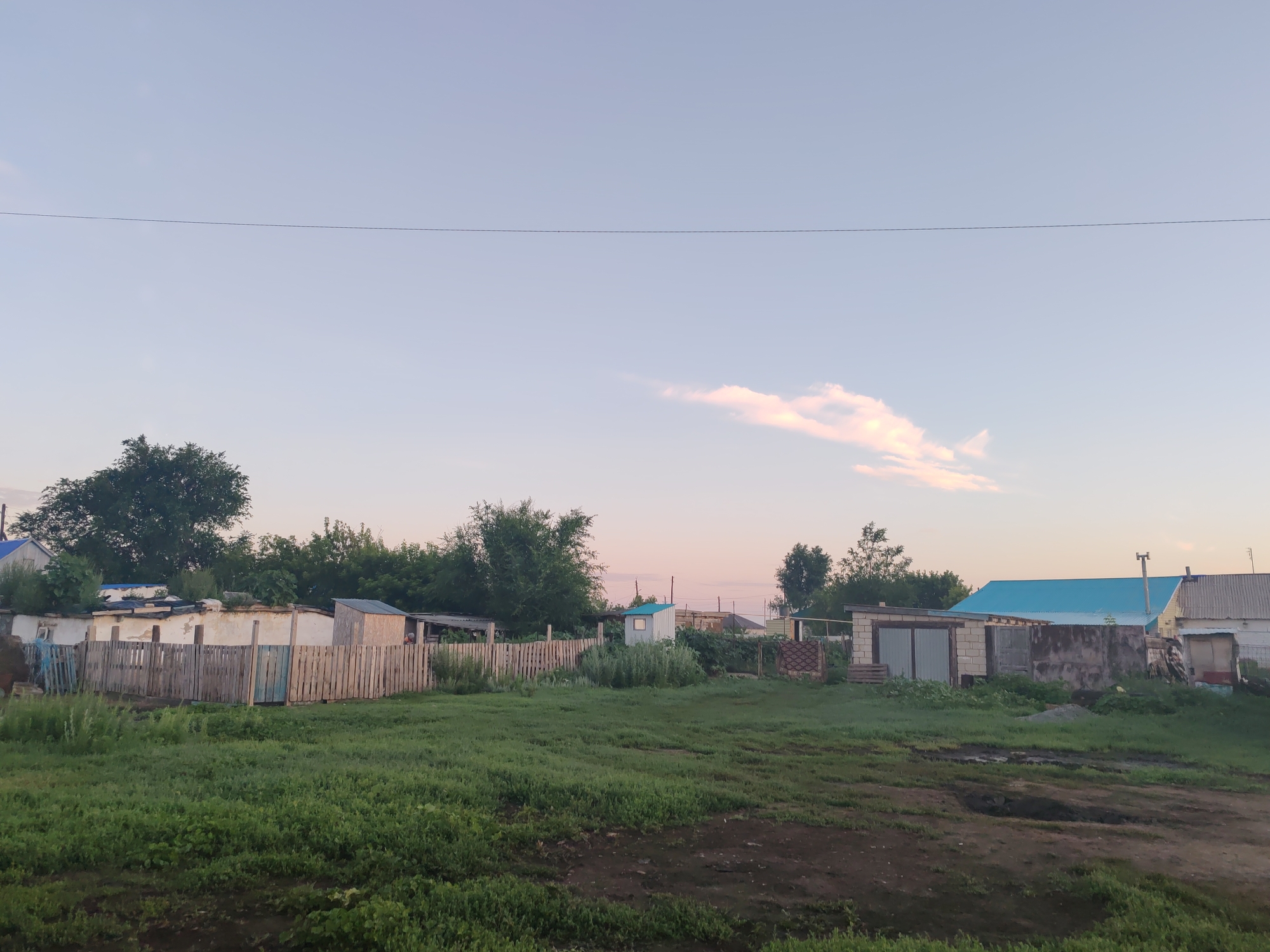 The image depicts a rural landscape, possibly on the outskirts of Akzhar, Kazakhstan. The scene is characterized by a vast, pale sky with a single elongated cloud illuminated by the setting sun. In the foreground, a green, somewhat overgrown grassy field stretches towards a rustic wooden fence. Beyond the fence, a collection of simple structures are visible, including small, weathered buildings and sheds with corrugated metal and blue roofs. The architecture suggests a modest residential or agricultural setting. Dense green trees form a backdrop to the structures, hinting at a natural, undeveloped environment. A thin wire stretches horizontally across the upper portion of the frame, adding a man-made element to the otherwise natural scene. The overall impression is one of a quiet, early evening in a rural community.