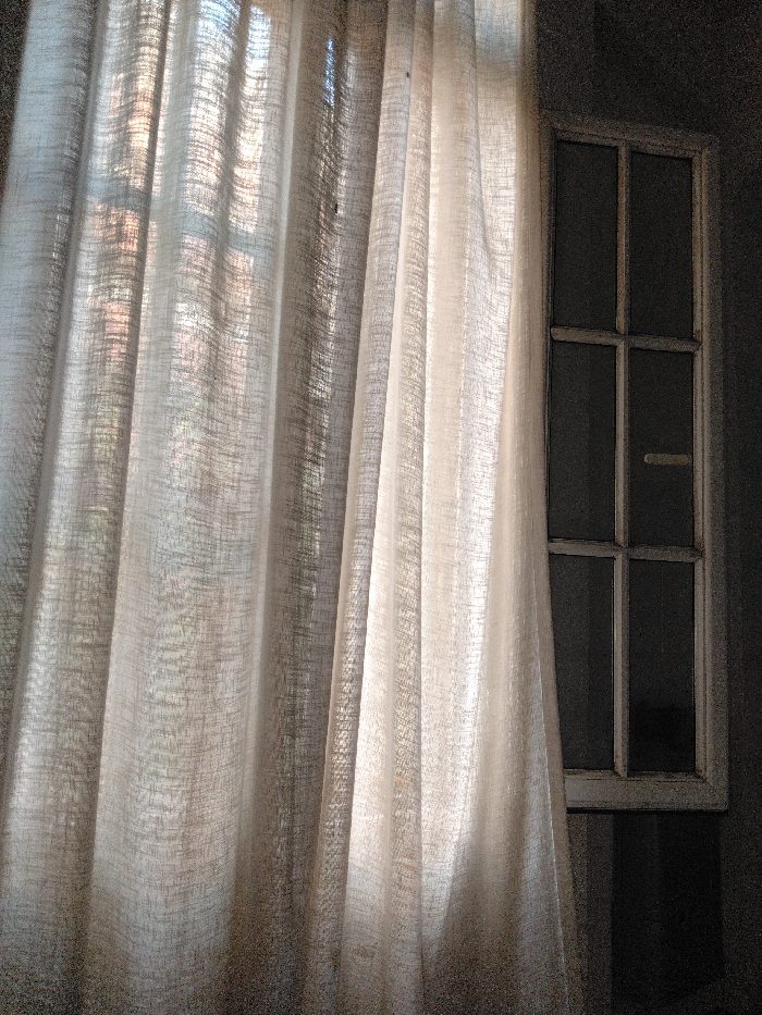 The image displays an indoor scene featuring a drawn, light-colored, semi-sheer curtain covering the majority of a window on the left side, extending approximately three-quarters across the frame. Sunlight filters through the curtain, creating bright vertical folds and shadows, revealing a subtle textured weave in the fabric. To the right, a multi-paned window with a light-colored frame is partially visible, showing at least six distinct panes arranged in a grid; a small, light-colored horizontal handle or latch is present on one of the central-right panes. The area surrounding the window on the right consists of a dark grey or shadowed interior wall. No people or discernible actions are present. The exterior view through the window is obscured by the curtain and window glass, appearing dark. The precise location within Buenos Aires, Argentina, cannot be determined from the visual evidence provided.