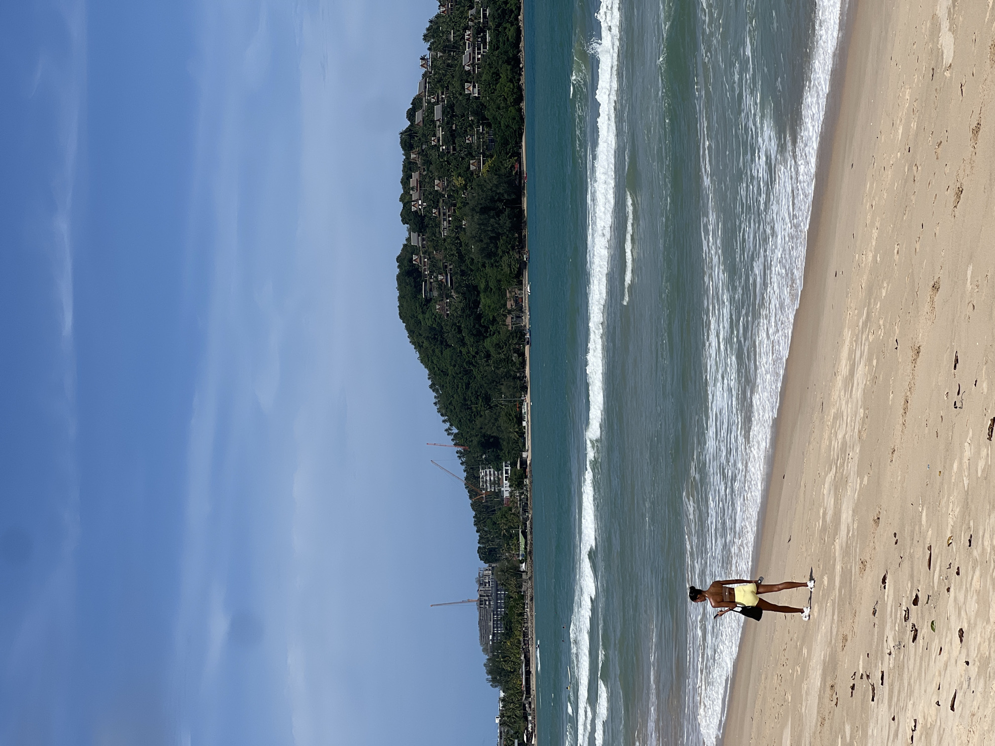 A full shot captures a person standing on a sandy beach, looking out at the ocean. The person is wearing yellow shorts and a yellow top, with white sneakers. A black bag is slung over their left shoulder, and they are holding a phone in their right hand. The beach is mostly empty, with a few scattered pieces of debris. In the background, the ocean stretches out to the horizon, with waves breaking on the shore. A lush, green hill covered in trees and buildings rises from the water in the distance. The sky is a clear blue with a few wispy clouds. Construction cranes are visible on the coastline, suggesting ongoing development.