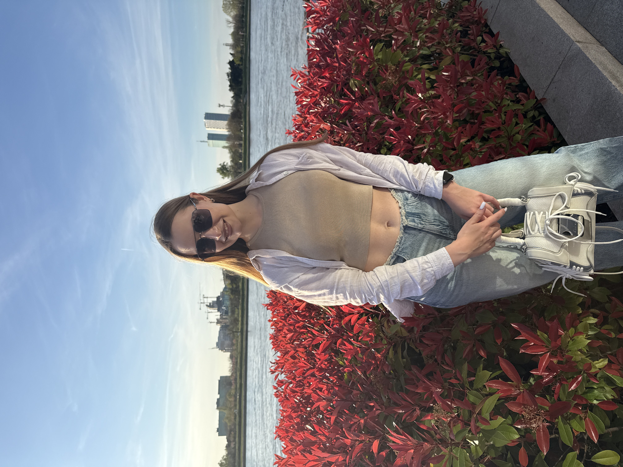 A young woman in sunglasses is seated, smiling, outdoors. She wears a light-colored tank top, a white button-down shirt, and blue jeans. She appears to be holding a white purse. Behind her is a vibrant shrub with red and green leaves. In the background, a body of water reflects a blue sky with distant city buildings. The location is in Belgrade, Serbia, identifiable by the skyline and the specific architectural style of the buildings in the distance.