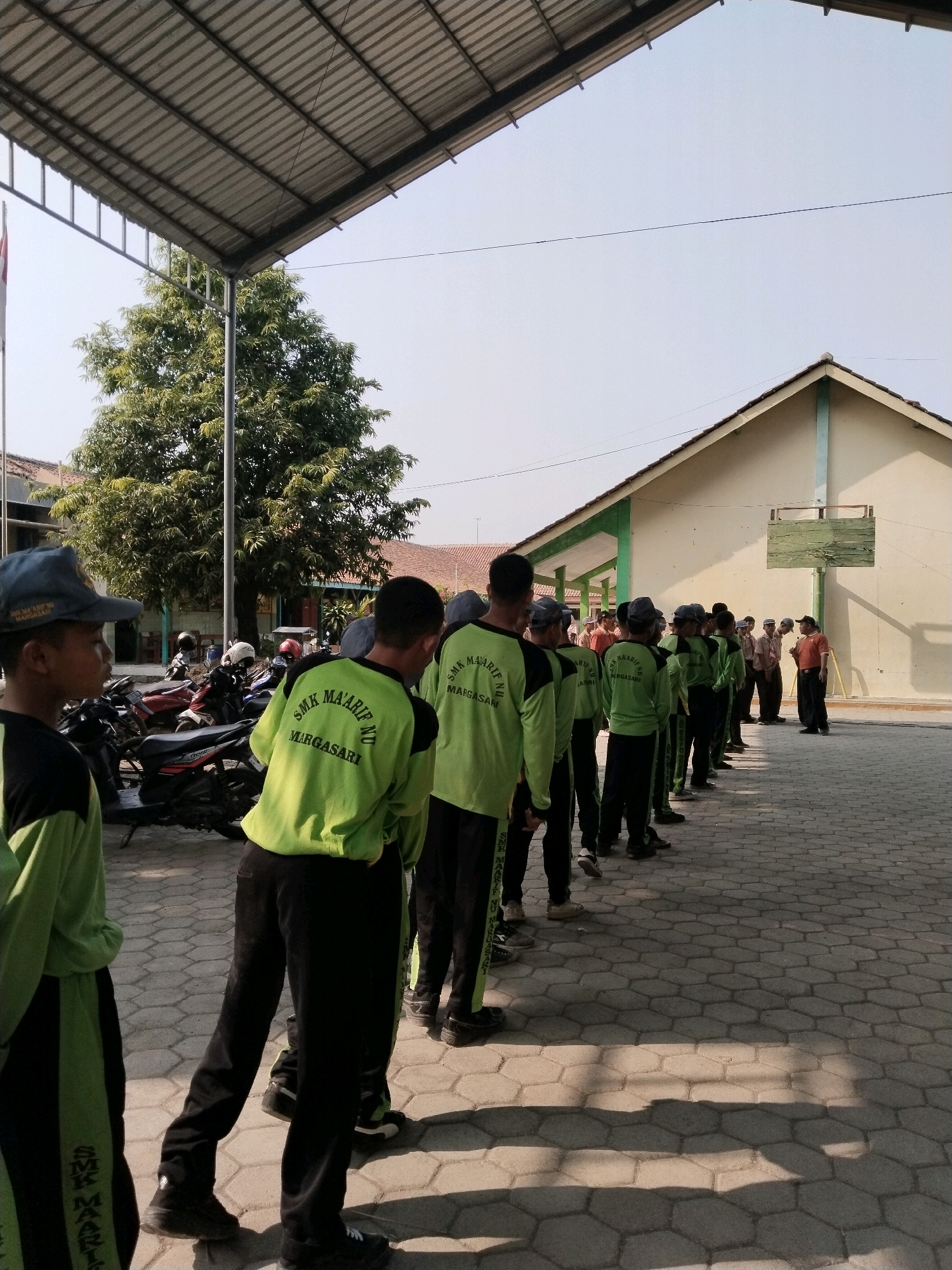 In Pakulaut, Indonesia, a group of young males in bright green long-sleeved shirts and black pants stand in a line outside a building. The shirts have the text "SMK MA'ARIF NU MARGASARI" printed on the back. The building behind them is light yellow with a green trim and a basketball hoop mounted on the wall. In the foreground, motorcycles are parked under a corrugated metal awning. The ground is paved with hexagonal stones. The sky is clear and blue. The scene appears to be an outdoor assembly or class activity.