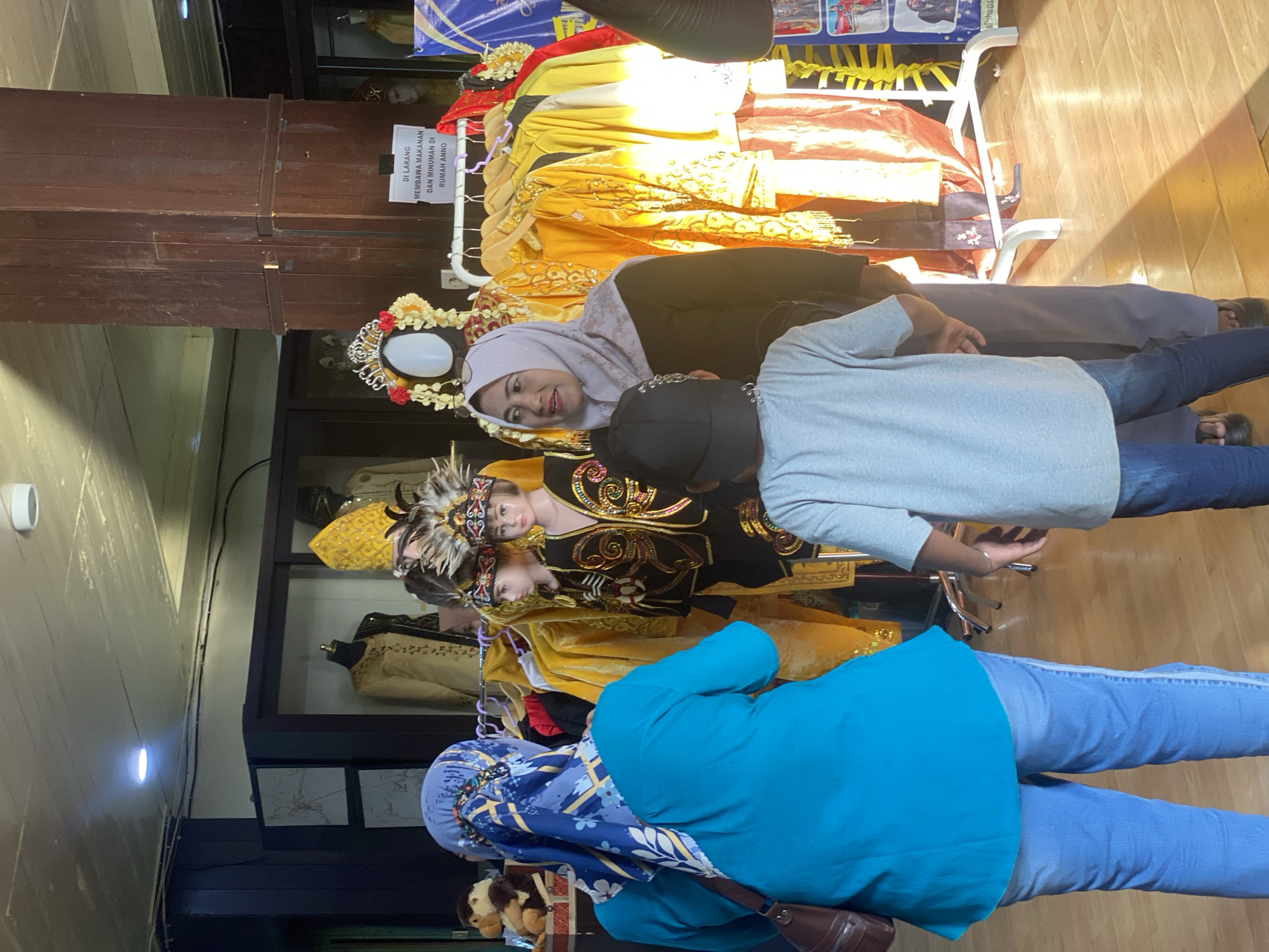 The image depicts the interior of a museum or cultural exhibition space in Banjarmasin, Indonesia. In the foreground, two individuals are visible from behind, a woman wearing a turquoise top and blue jeans with a blue and white patterned headscarf, and a young person wearing a grey t-shirt and a black baseball cap. To their right, a woman in a light purple hijab and dark long-sleeved shirt is looking to her left. In the mid-ground, several mannequins are dressed in traditional attire. One mannequin, to the left of the woman in the hijab, is wearing a black and gold outfit with a feathered headdress. Another mannequin, to the right, is adorned with a crown and flowers. A rack of brightly colored traditional garments, primarily yellow and orange, is displayed behind the mannequins. A wooden pillar stands prominently in the mid-right of the frame, with a sign attached to it in Indonesian that reads "DI LARANG MEMBAWA MAKANAN DAN MINUMAN DI RUMAH ANNO" (Prohibited to bring food and drinks in the Anno House). The floor is made of polished wood. In the background, glass display cases contain more traditional clothing, and a large wooden structure, possibly part of the building's architecture, is visible. The lighting suggests an indoor environment with artificial illumination.