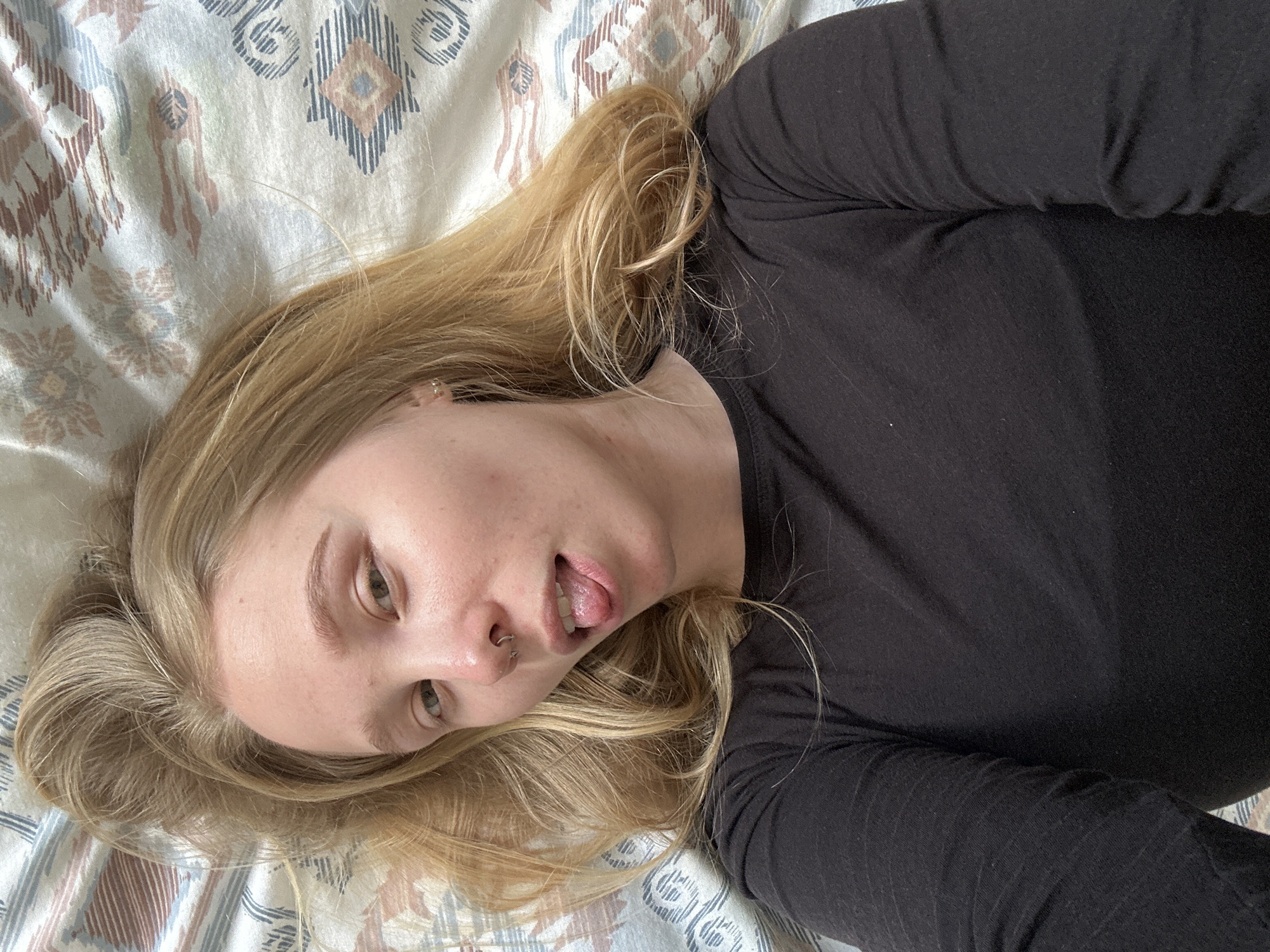 A person with long, wavy blonde hair lies on their back, looking slightly to the viewer's left. They are wearing a black long-sleeved top. The individual has a septum piercing, visible as a silver horseshoe-shaped ring, and a small silver hoop piercing in their right ear. Their light-colored eyes are open, and their tongue is playfully sticking out slightly. The background is a patterned bedsheet or blanket featuring geometric and abstract designs in muted tones of light blue, beige, and brown on an off-white fabric. The lighting suggests it is daytime. The overall impression is casual and relaxed. There is no visible text in the image. The provided location context for this image is Budva, Montenegro.