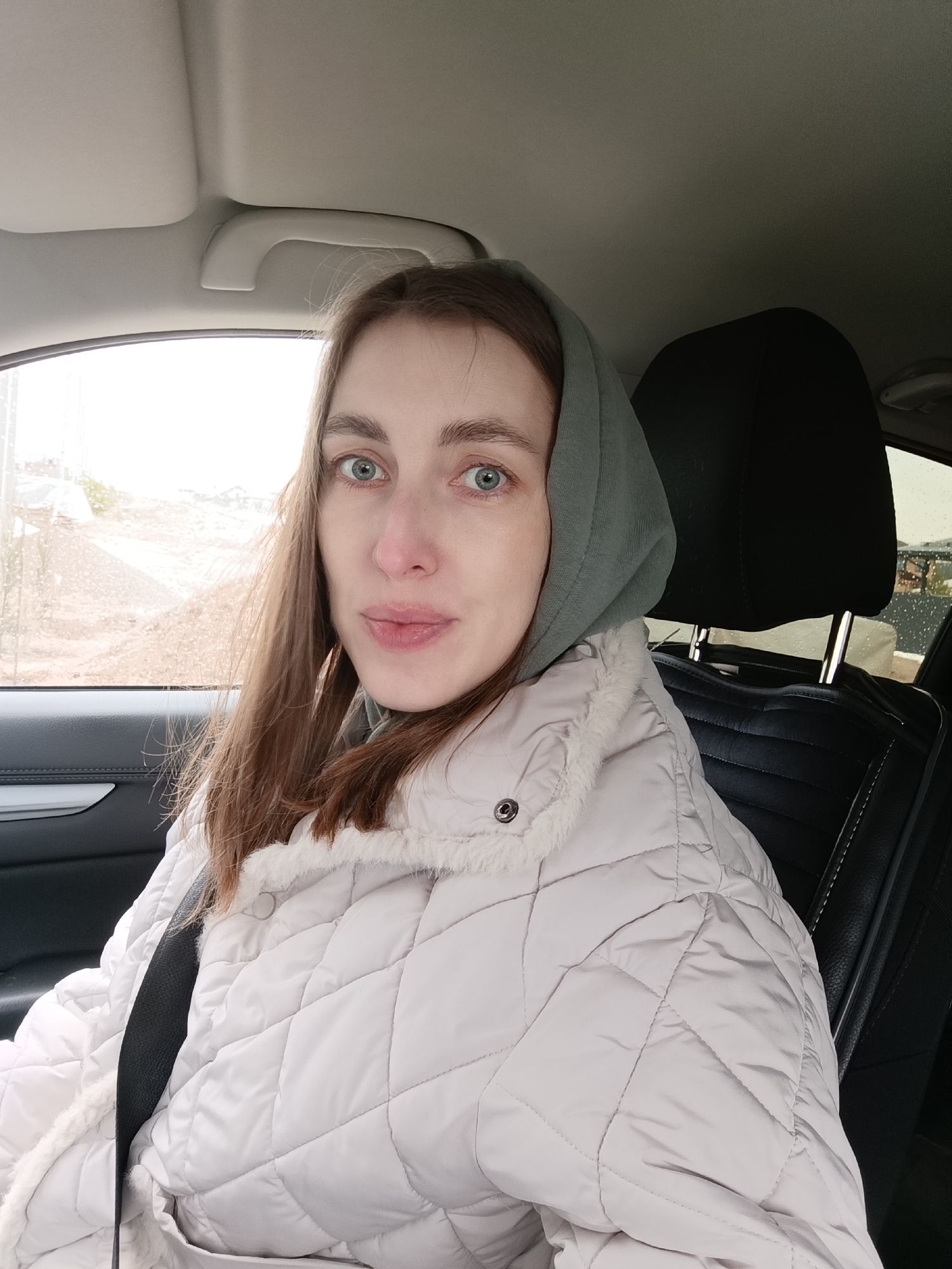 A woman is sitting in the driver's seat of a car. She is wearing a light-colored quilted jacket with fur trim and a grey hoodie. The seatbelt is fastened across her chest. Outside the car window, there is a view of a dirt road and some buildings in the distance. The location is likely in Chaikovsky, Russia. It is possibly a rainy day as there appear to be raindrops on the window behind the headrest.