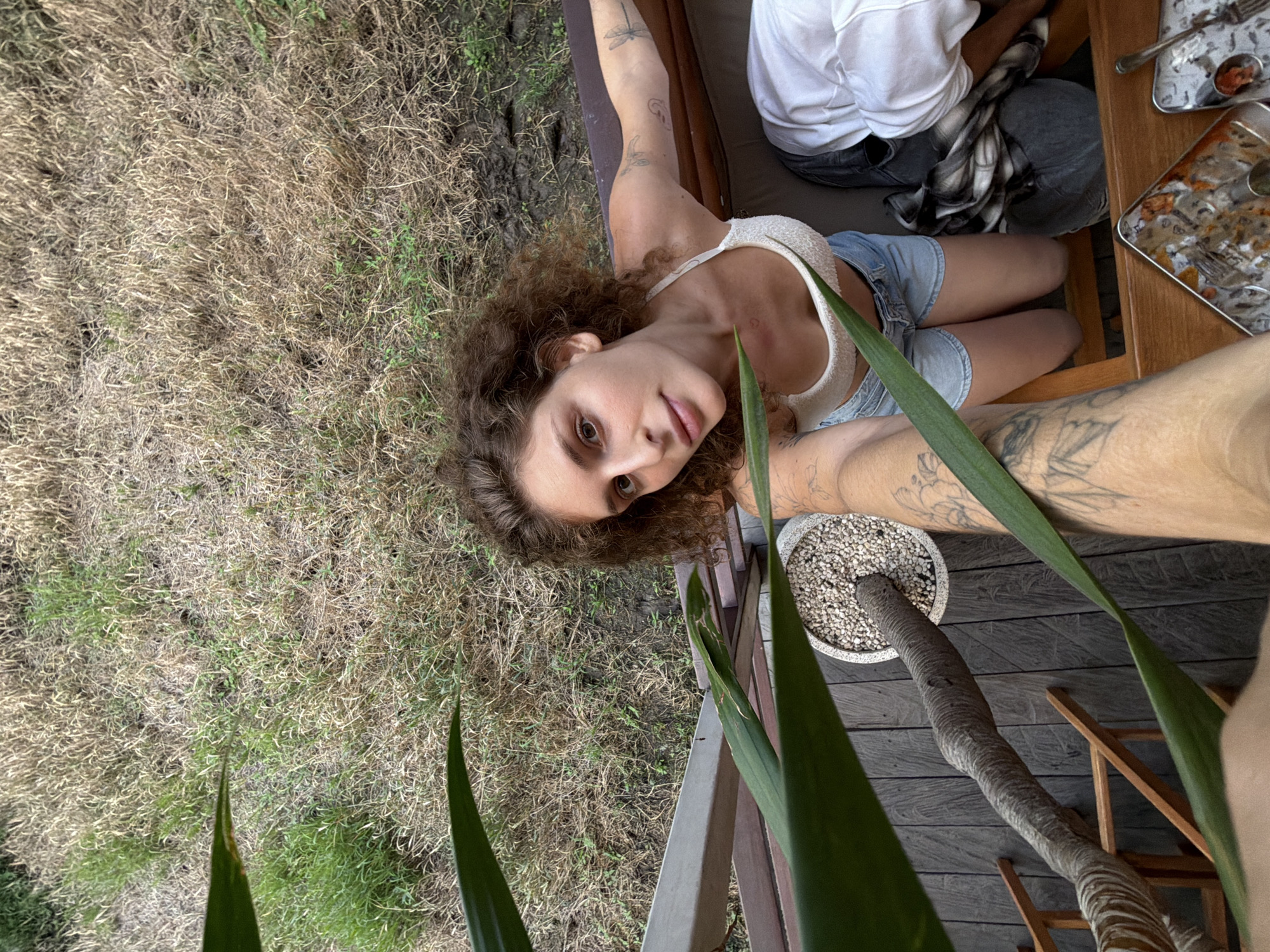 A young woman with curly brown hair, wearing a white bikini top and denim shorts, takes a selfie from a high angle on a wooden deck. She is looking up at the camera. Large green leaves are in the foreground. In the background, dry grass and some green patches are visible. A potted plant with small stones is on the deck. Part of another person wearing a white shirt and jeans is visible to the right. In the bottom right corner, two metal trays with remnants of food are present. The scene appears to be outdoors in a natural, somewhat unkempt setting, possibly at a resort or private residence in North Kuta, Indonesia.
