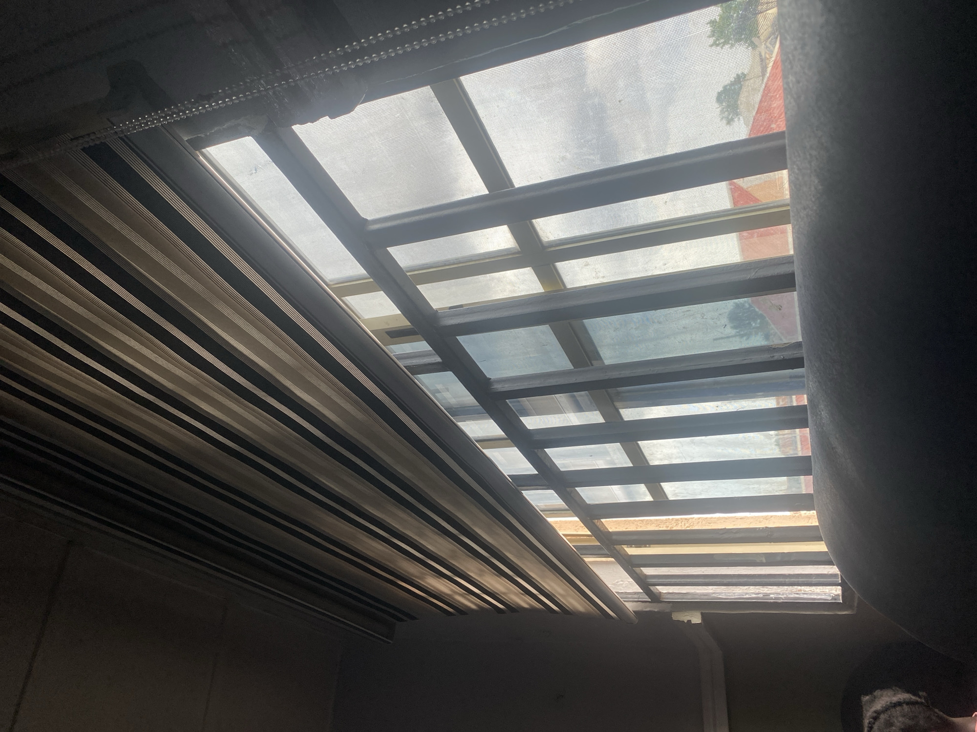This image captures a view through a window, looking outward over a residential area in Jos, Nigeria. The window is fitted with a roller blind that has alternating opaque and sheer horizontal stripes, allowing diffused light to enter. Vertical bars, likely part of a security grate, are visible across the lower portion of the window. Beyond the bars, rooftops of houses, some with red-tiled roofs, and scattered trees are discernible under a cloudy sky. In the foreground, a dark, upholstered surface, possibly a couch or chair, occupies the bottom of the frame, with a hint of a person's dark hair visible on the far left. The overall lighting suggests it is daytime, with natural light filtering through the window.