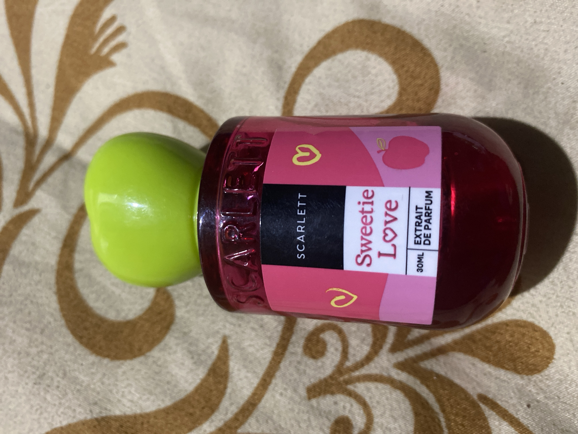 The image shows a bottle of "Sweetie Love" perfume from the brand Scarlett. The bottle is red and has a pink label with the text "Sweetie Love" and "EXTRAIT DE PARFUM". The cap of the bottle is shaped like a green apple. The bottle is placed on a patterned fabric that appears to be a blanket or tablecloth. The background is blurred and shows the same pattern as the fabric. There are no people or any other objects visible in the image. The location in Ta', Indonesia is not discernible from the image.