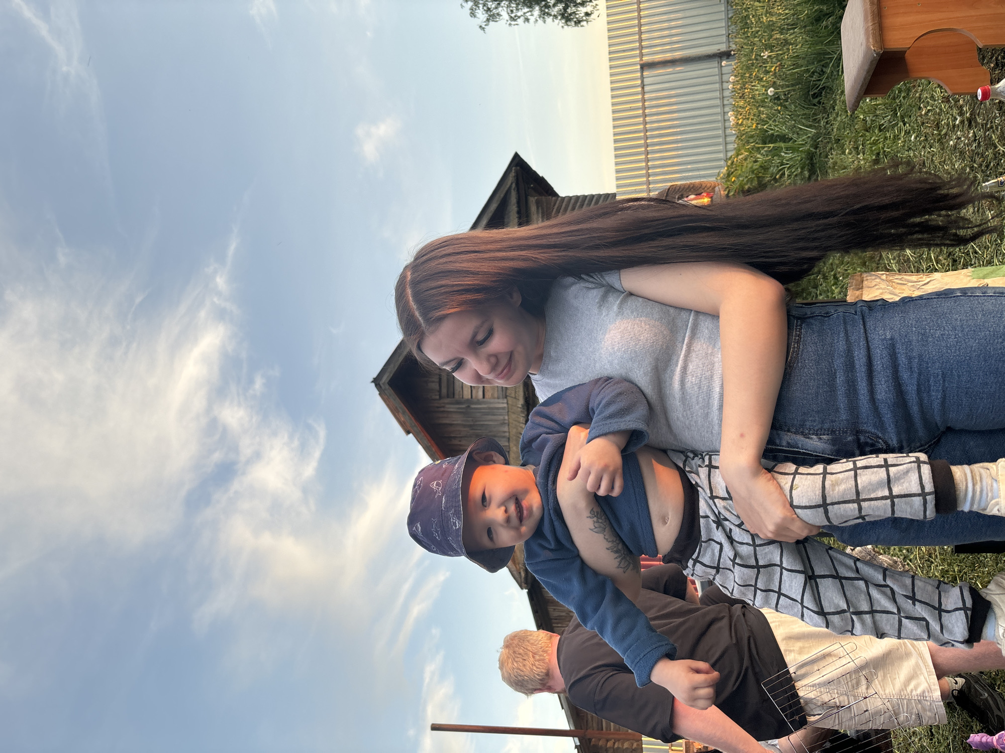 A woman with long, dark hair is holding a young child wearing a blue hat and navy shirt. They are outside, with a partially visible wooden building behind them. The sky is light blue with wispy clouds. A man with blond hair in a black shirt is also partially visible. Additional objects in the scene include a metal grill, green grass and a metal fence. The location is likely the village of Фоки, Russia based on the location.
