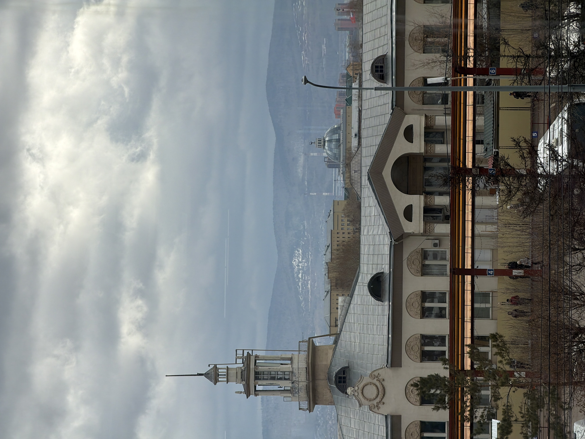 This image captures an urban scene in Krasnoyarsk, Russia, set against a backdrop of hazy, snow-dusted mountains under an overcast sky. The photograph appears to be taken on a cloudy day, with a soft, diffused light, and the bare trees suggest late autumn or winter.
In the foreground and midground, several ornate buildings with classical or historic architectural styles dominate. The most prominent structure features a light-colored facade, possibly beige or pale yellow, adorned with arched windows, decorative moldings, and circular embellishments. Its dark, tiled roof is punctuated by several dormer windows, some round and some arched. A distinctive multi-tiered tower or spire rises from the building, culminating in a pointed top, adding a prominent vertical element to the skyline. Another building with a similar architectural character is visible further back.
At street level, below the buildings, a public space or street is discernible. Bare deciduous trees line the area, and several people are scattered throughout, suggesting a typical daytime activity. There are structures that resemble public transport stops, featuring canopies and supporting posts.
The background is defined by a range of blue-gray mountains, their slopes showing patches of snow or ice. Amidst the distant urban sprawl at the base of these mountains, a modern-looking domed building stands out. A faint contrail from an aircraft is visible in the cloudy sky.
Visible text includes the numbers "9" on a red vertical signpost, "4" on another similar red sign, and "Б 5" (likely a bus or tram route indicator) on a blue sign.