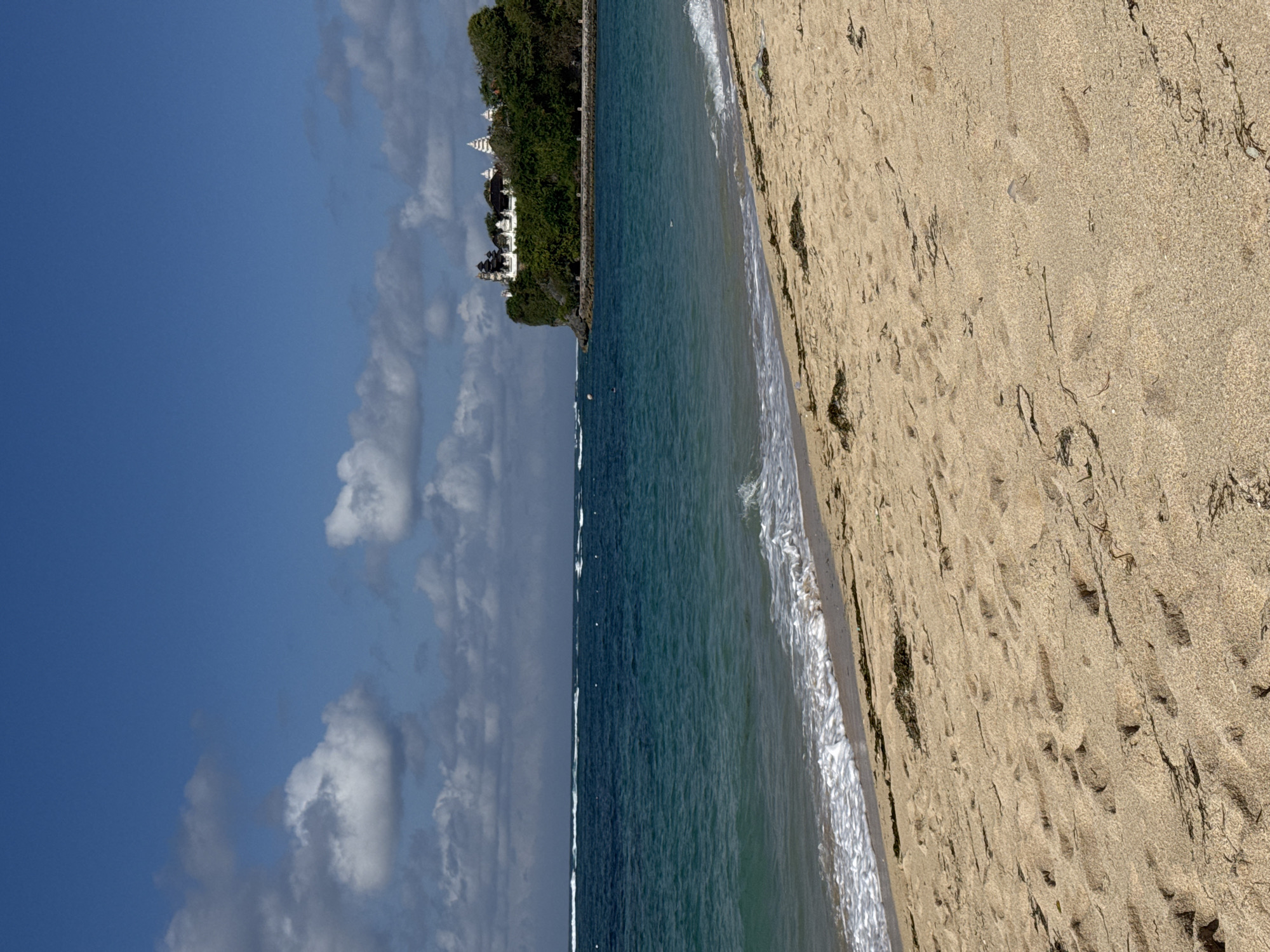 The image displays a coastal landscape, oriented as if taken from a beach looking out towards the ocean and a distant landmass.
In the foreground, a light brown sandy beach stretches along the right side, showing numerous footprints and scattered dark strands of seaweed or debris. Gentle white-capped waves break softly on the shore, marking the transition from sand to sea.
The middle ground is dominated by the calm, blue-green waters of the ocean. Small white foamy lines indicate waves further out, and a few indistinct dark spots, possibly small objects or distant swimmers, can be seen on the water's surface.
In the background, a verdant, tree-covered cliff or hill rises from the ocean. Perched atop this green expanse are several white or light-colored buildings with distinct, multi-tiered rooflines, suggestive of traditional architecture, possibly temples or resort structures. A dark wall or barrier runs horizontally along the edge of the cliff, separating the foliage from the sea.
Above the distant land and stretching across the left side of the frame is a bright blue sky, adorned with scattered white, puffy clouds.
The scene depicts a clear, sunny day, likely mid-day, evoking a sense of tranquility and a tropical setting. No people are distinctly visible, and there is no discernible text.