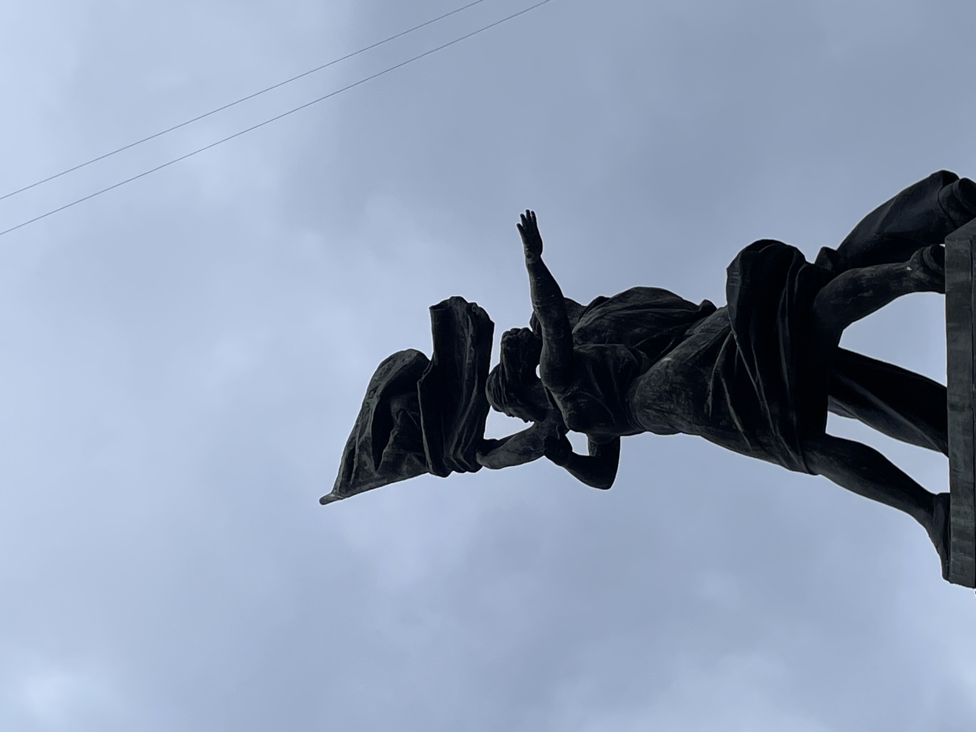 The image shows a low-angle view of a dark-colored statue against a cloudy sky. The statue depicts two figures, one with an arm raised and the other holding a flag. A thin wire is visible in the upper left corner of the frame. The statue is the monument "Keyboard," a sculpture located in Yekaterinburg, Russia.