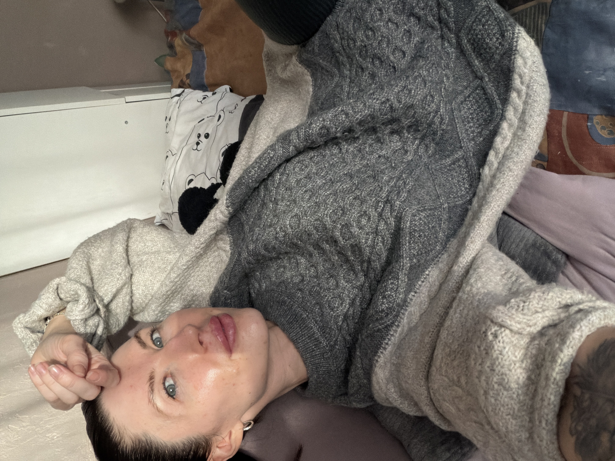 The image presents a close-up, vertical shot of a person, identified as a woman, lying down and looking upwards towards the camera. She has dark hair, striking blue eyes, and a neutral to slightly pouting expression with her lips lightly parted. A small silver hoop earring is visible in her left ear. She is wearing a cozy, thick knitted sweater or cardigan that features two tones of grey: a darker grey section with prominent cable knit patterns, and a lighter grey section, possibly layered or integrated into the design. Intricate dark tattoos are visible on her left arm, extending from beneath the sweater's sleeve.
The setting appears to be indoors, likely a bedroom or living area. Behind her head, a white pillow with multiple black and white cartoon bear faces adds a whimsical touch. To the left of the pillow is a plain white piece of furniture, possibly a headboard or a storage unit. Surrounding her are various bedding items, including a light purple or pinkish fabric underneath her and a multi-colored patterned fabric further in the background, suggesting a comfortable and casual environment. The image is a relaxed portrait, possibly a selfie, capturing a quiet moment. No visible text is present in the image. The location context for this scene is Perm, Russia.