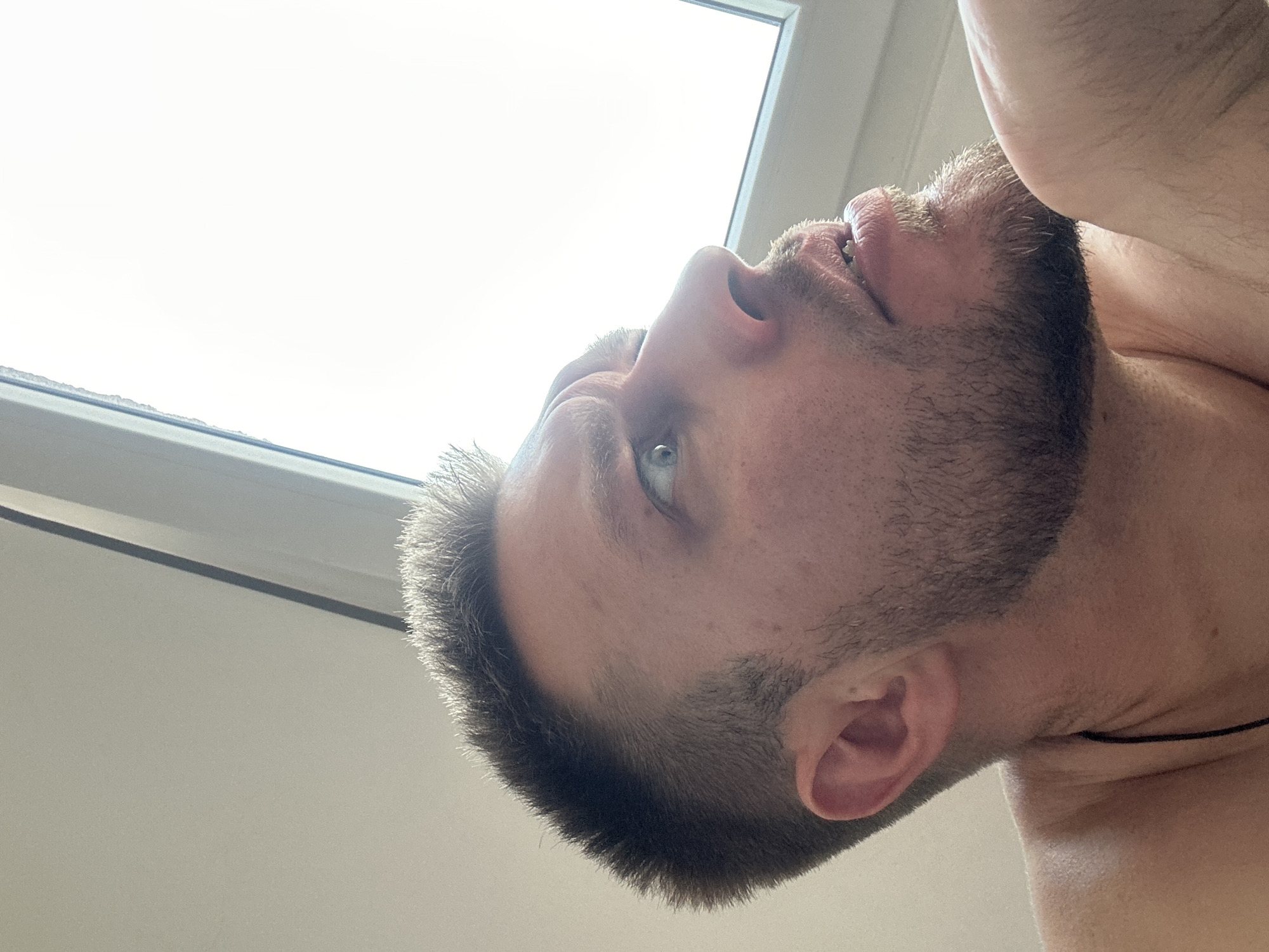 In the city of Zvezdara Urban Municipality, Serbia, a close-up, low-angle shot captures a man looking upwards and to the right. The man has a light complexion, short brown hair with a textured cut, and a light stubble beard. His right eye is clearly visible, with a surprised or curious expression indicated by raised eyebrows. His mouth is slightly open, with his lower teeth visible. The background consists of a bright, overcast sky seen through a window frame, with the right edge of the frame partially visible. The man is shirtless, and a thin black cord is visible around his neck.